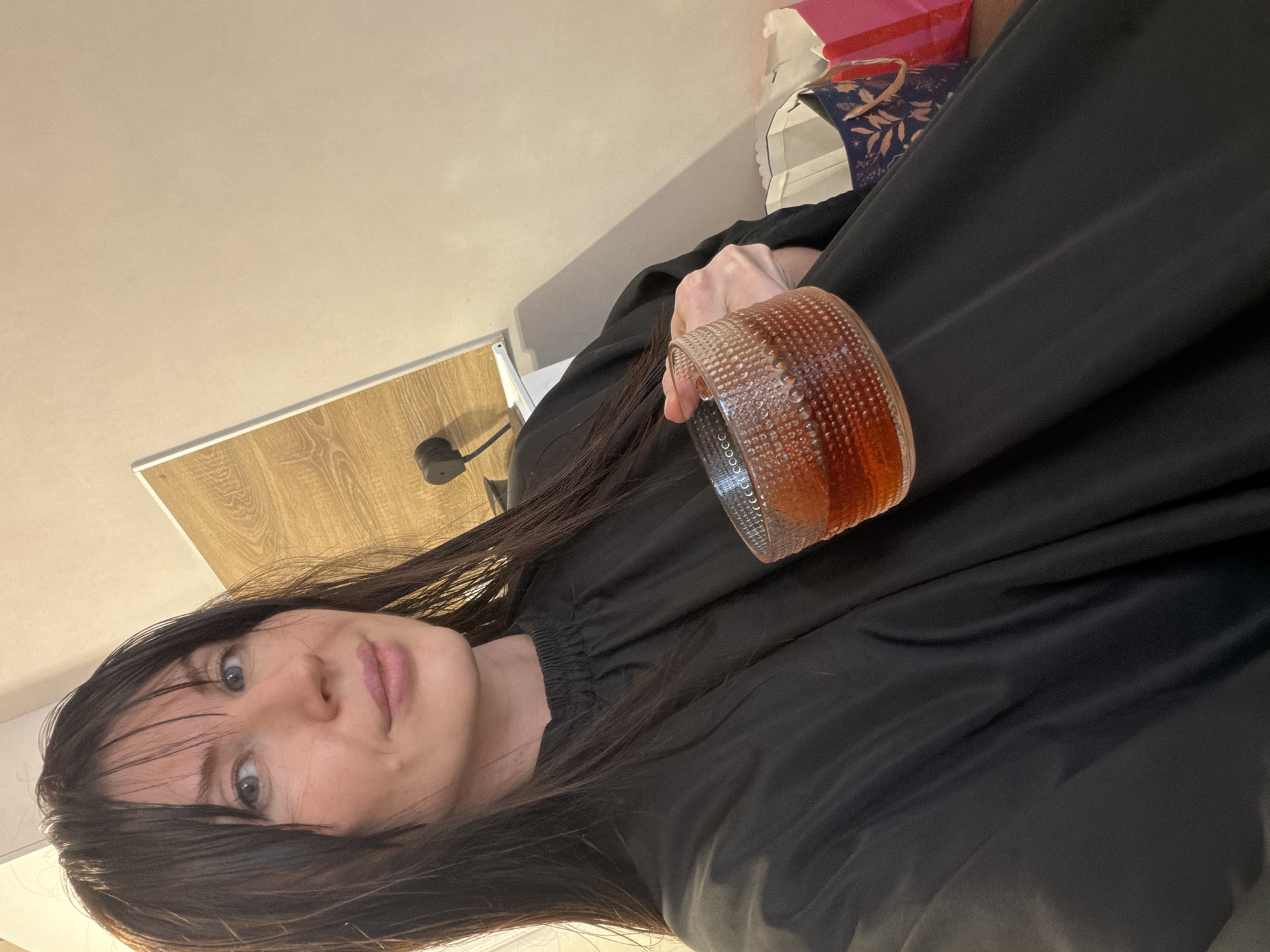 A woman with dark hair and bangs is holding a glass of amber-colored liquid. She's wearing a black garment, possibly a salon cape. The background includes a light-colored wall, a section of wood-grain paneling, and a partially visible shopping bag. The location is not discernible from the image.