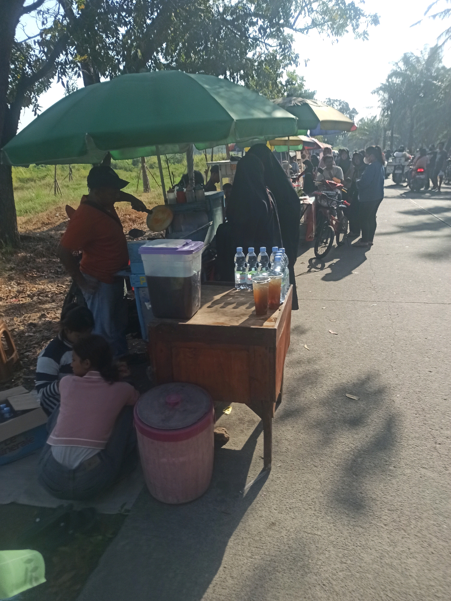 The image depicts an outdoor scene in Telagamurni, Indonesia, characterized by a street-side vendor operation. A vendor, wearing an orange shirt and a dark cap, is cooking or preparing food on a flat surface, possibly a griddle, using a utensil. He is positioned under a large green umbrella, which also shelters a makeshift food cart or stall. To the right of the vendor, a wooden table holds several bottles of water and two plastic cups filled with a brown beverage, presumably iced tea or coffee, along with a large clear container filled with the same liquid.
In the foreground, to the left of the vendor, two individuals are seated on the ground, facing away from the camera. One is wearing a striped long-sleeved shirt and a dark head covering, while the other wears a pink long-sleeved shirt and dark pants. A large pink plastic bin with a red lid is situated near them.
In the background, a number of people are visible, some standing near other stalls or umbrellas of varying colors (yellow, red, and blue). Several motorcycles and scooters are parked or moving along the asphalt road, indicating a public thoroughfare. The environment includes trees with dense foliage, casting shadows on the ground, suggesting it is daytime. The overall impression is a busy, informal commercial area by a road.