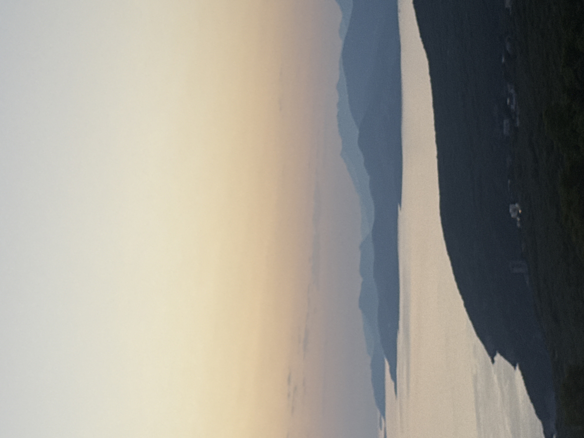 The image captures a serene coastal landscape at dusk, likely in or around Kovačko Polje, Montenegro. In the foreground, a dark, forested hillside slopes down towards the water, with a few indistinct buildings or structures visible amidst the trees. The middle ground features a calm body of water reflecting the soft, pastel hues of the sky. Distant, layered mountain ranges, silhouetted against the fading light, form the background. The sky transitions from a pale yellow near the horizon to a soft blue at the top, with subtle hints of clouds. The overall atmosphere is tranquil and peaceful, characteristic of a sunset over a Mediterranean coastline.