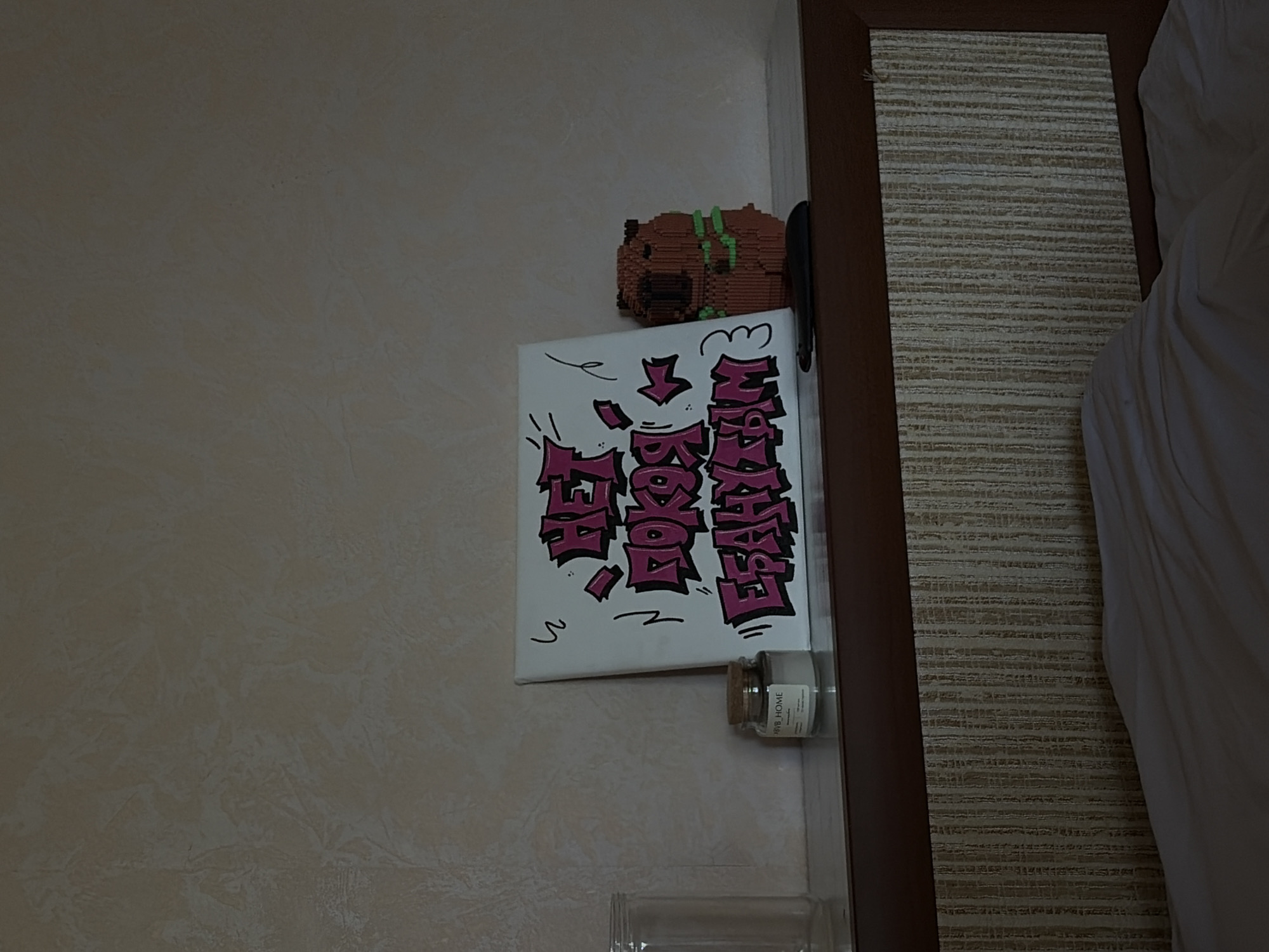 The image shows a portion of a wall in what appears to be an interior space. The wall is painted in a light, neutral color. Attached to the wall is a white canvas with pink graffiti-style writing on it. Above the canvas is a small, blocky, brown object. Below the canvas is a small, clear glass jar with a cork lid and a label that reads "MRB HOME". There is also a clear, plastic container partially visible at the bottom of the frame. To the right of the wall, there is a brown, wooden frame with a beige, textured panel behind it, as well as a white sheet or fabric. The location is Chaikovsky, Russia.
