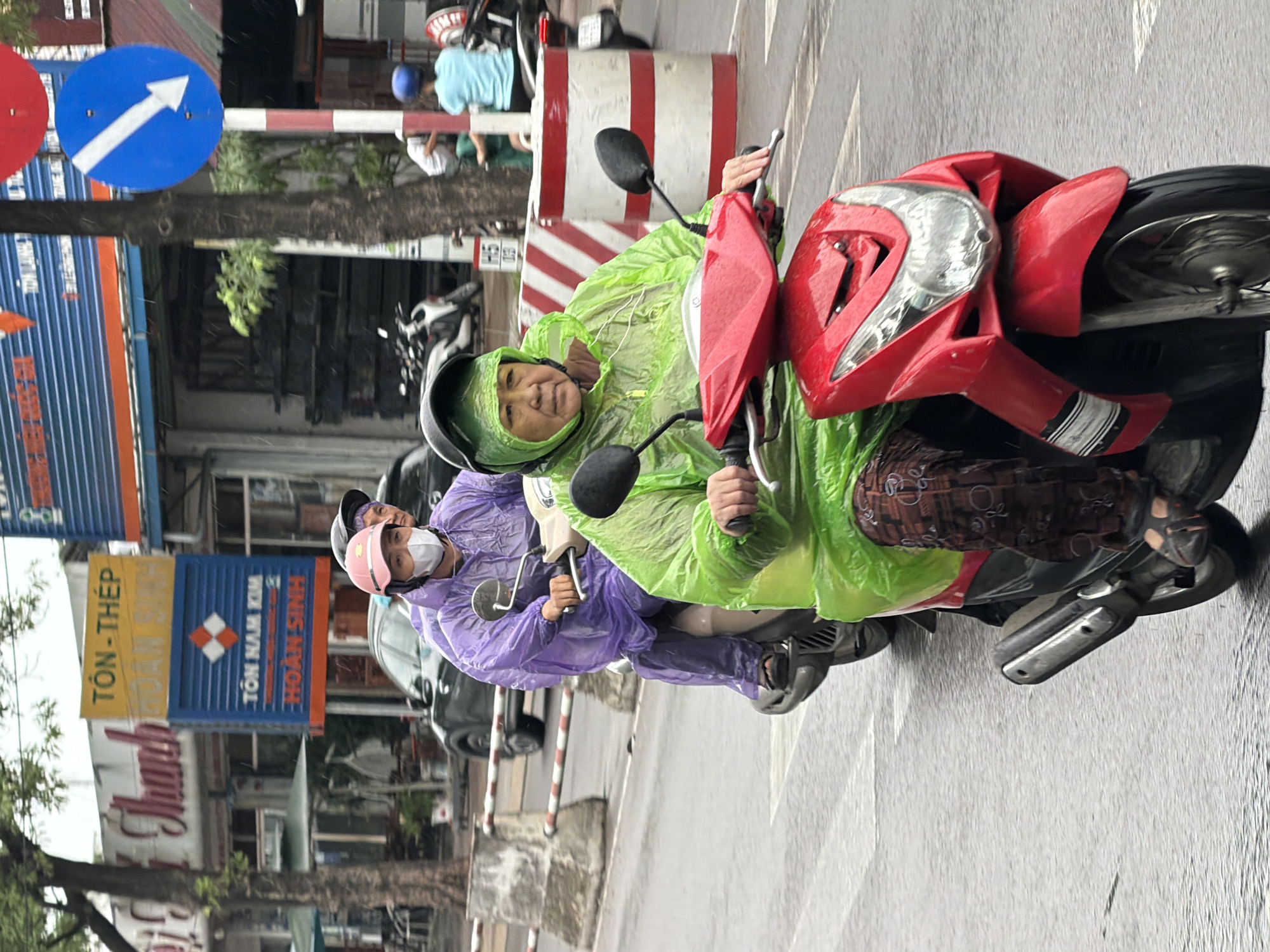 Two individuals are riding a red scooter in Nha Trang, Vietnam during what appears to be rainy weather. The person in the front is wearing a bright green rain poncho and a dark helmet, and the person behind them is wearing a purple rain poncho and a pink helmet, with a white face mask. The scooter has a red body and is traveling on a wet, paved road. In the background, there are buildings with signs, including one with Vietnamese text and another with a blue and white directional arrow. Several other scooters and a car are also visible in the background, along with trees and barriers along the side of the road.