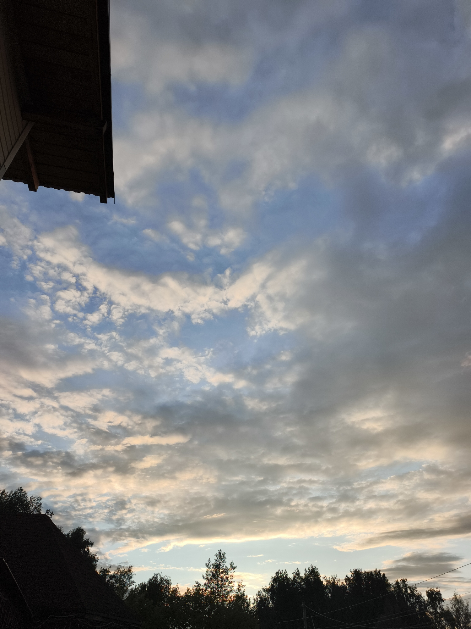 The image captures a dusky sky with scattered clouds, tinged with hues of pink and orange from the setting sun. A dark, silhouetted roofline from a building extends from the upper left corner, and another roofline, along with the tops of dark green trees, forms a silhouette along the bottom of the frame. The overall atmosphere is calm and serene, suggesting the end of a day. The scene is set in an outdoor environment, likely a residential area, given the presence of rooftops and trees. There are no people or animals visible in the image.