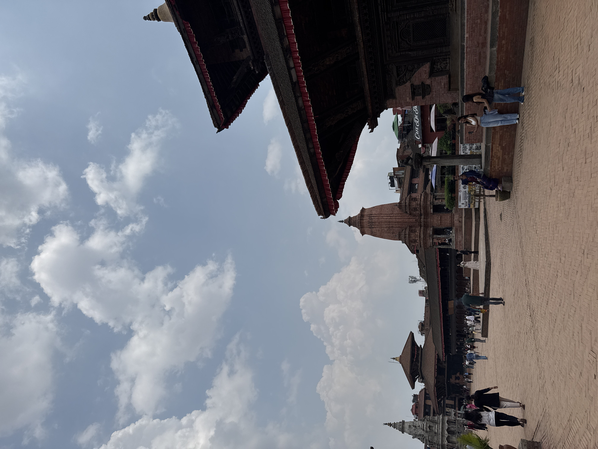 The image shows a scene in Bhaktapur, Nepal, likely in Durbar Square. Several traditional wooden buildings with multiple tiered roofs are visible, constructed with intricate carvings. A brick-paved square is filled with people; some are walking, while others stand or sit. A tall brick stupa rises in the background, and the sky is filled with clouds. A "Cafe De Silk" sign is visible behind one of the buildings.