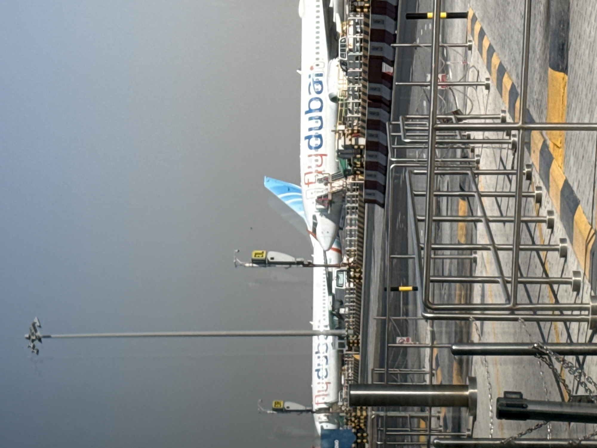 The image captures a scene at Dubai International Airport (DXB), United Arab Emirates. Two Flydubai airplanes are visible parked at the gate. The aircraft are predominantly white with the airline's blue and red logo on the fuselage and tail. In the foreground, metal railings and barriers line the walkway, with yellow and black striped speed bumps on the tarmac. Tall airport lighting poles with multiple lamps are also present, casting light on the scene. The sky is clear and a pale blue. The scene appears to be during daylight hours, with the sun creating some glare on the aircraft. There are no people clearly visible in the image.