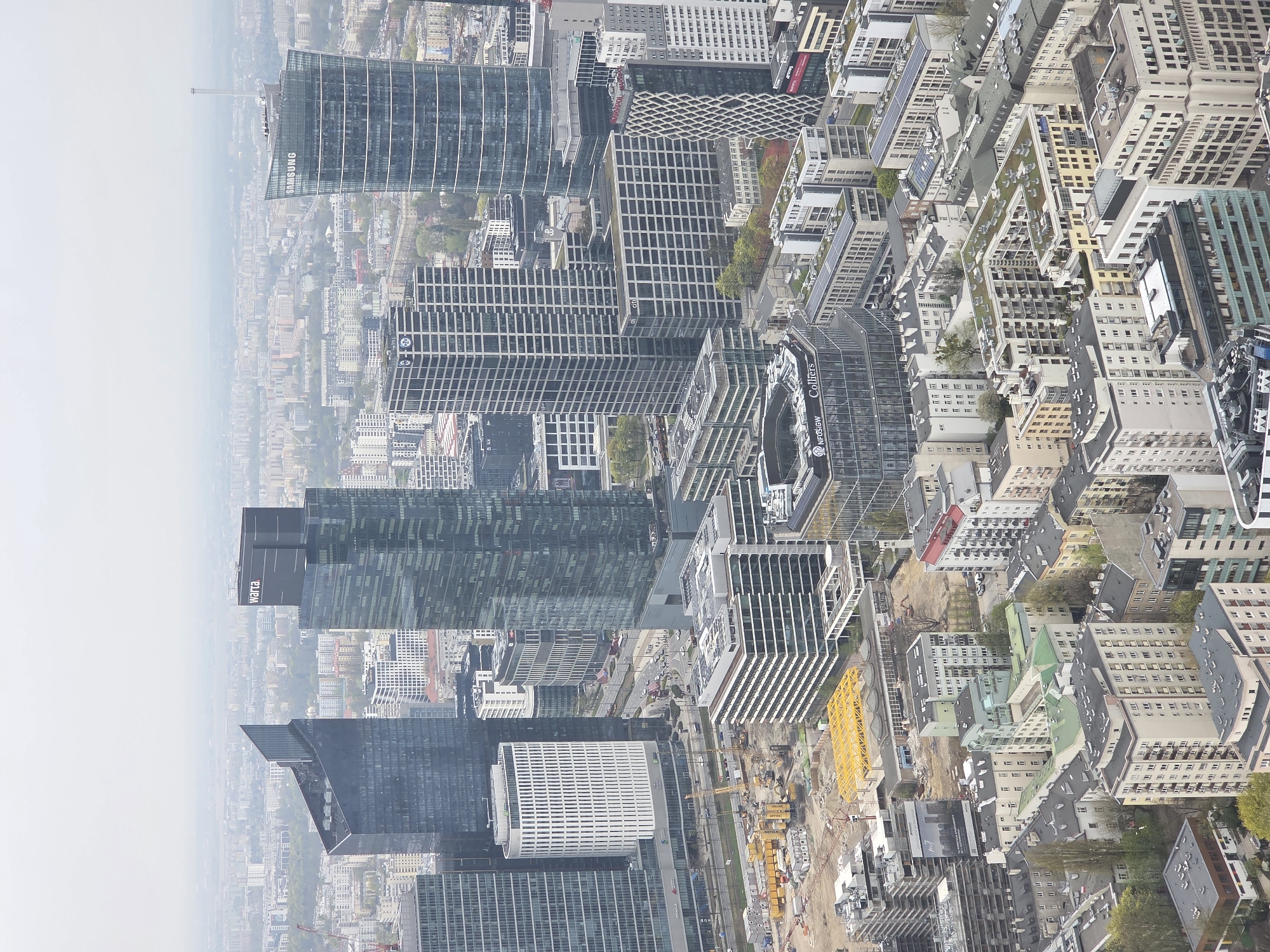 The image shows an aerial view of Warsaw, Poland, dominated by modern skyscrapers and interspersed with older buildings. Skyscrapers with glass facades form the core of the cityscape. A large construction site with yellow cranes and heavy equipment is visible amidst these buildings. The environment is hazy, particularly in the distance. There are no people explicitly visible.