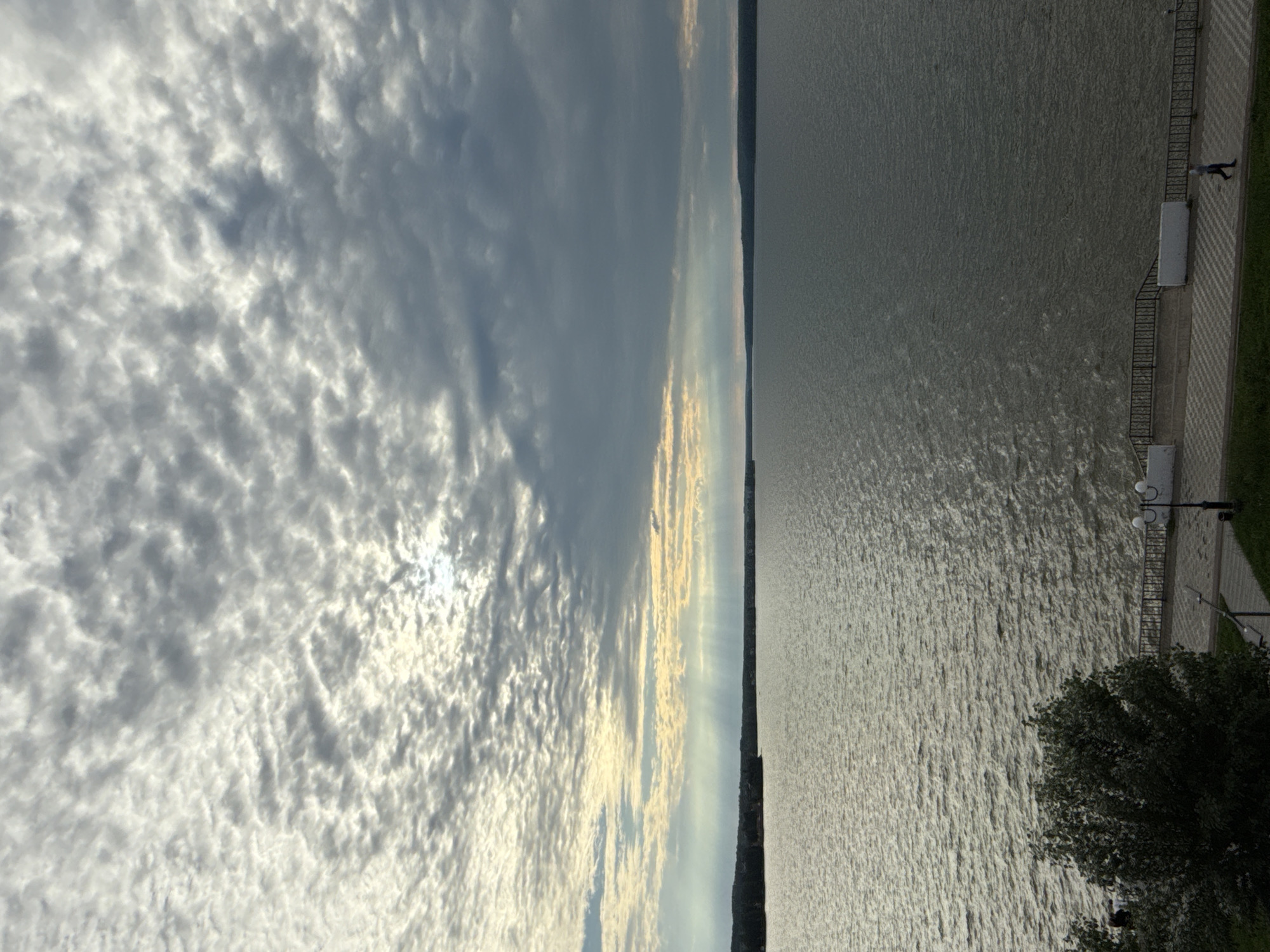The image depicts a wide, tranquil body of water, likely a reservoir or lake, adjacent to a cityscape. The sky is predominantly overcast with textured, mottled clouds, but shafts of sunlight break through, creating a dramatic effect above the horizon. A distant treeline marks the opposite shore, creating a sharp contrast with the vast expanse of water. In the foreground, a paved walkway runs along the water's edge, bordered by a dark railing and decorative white structures. A lone figure, dressed in dark clothing, walks along the pathway. Trees and greenery are visible on the left side of the walkway, suggesting a park or embankment area. The overall atmosphere is serene, with a hint of melancholic beauty due to the cloudy sky. This scene is identifiable as the embankment of the Votkinsk Reservoir in Votkinsk, Russia.