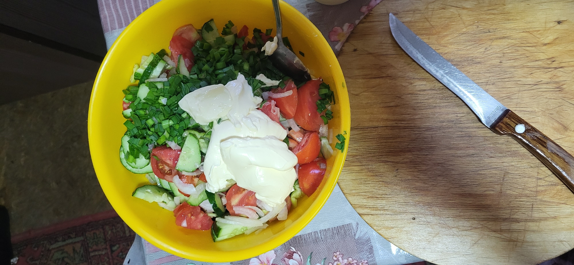 A high-angle medium shot captures a bright yellow bowl filled with a freshly prepared salad. The salad consists of chopped tomatoes, sliced cucumbers, sliced onions, and finely chopped green onions, topped with dollops of a creamy white dressing, likely mayonnaise. A silver spoon is partially submerged in the salad, suggesting it is being served or mixed.
The bowl sits on a patterned tablecloth with floral motifs. In the foreground, a round wooden cutting board displays a knife with a wooden handle and a stainless steel blade. The blade has the inscription "ROSTFREI" visible. The background shows a dimly lit surface and part of a patterned rug. The scene appears to be indoors, likely in a kitchen or dining area, and the style of the food and presentation suggests a home setting. Based on the provided context, this image is located in the city of Oskemen, Kazakhstan.