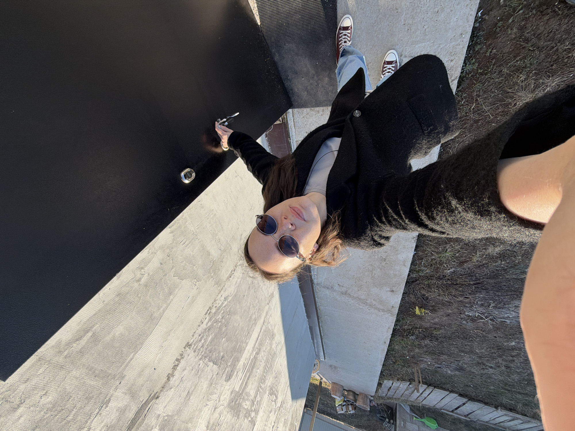 A young woman captures a selfie outdoors in Chaikovsky, Russia. She is positioned with a dark, textured vertical surface to her left, contrasting with a light-colored concrete ground extending to her right and beneath her feet. The woman has long brown hair and wears round, dark sunglasses. She is dressed in a light gray top, a long, dark (likely black) textured coat or cardigan, and light blue jeans. Her feet are visible, wearing burgundy high-top sneakers with white laces and soles. Her right arm is extended, holding a small set of keys or a keychain, likely operating the camera for the selfie. The strong, bright daylight casts clear shadows, indicating sunny weather. Beyond the concrete, a patch of dry grass and dirt is visible, along with some scattered debris, pots, and a green plastic toy in the background. The woman's expression is neutral, partially obscured by her sunglasses. No discernible text is visible in the image.