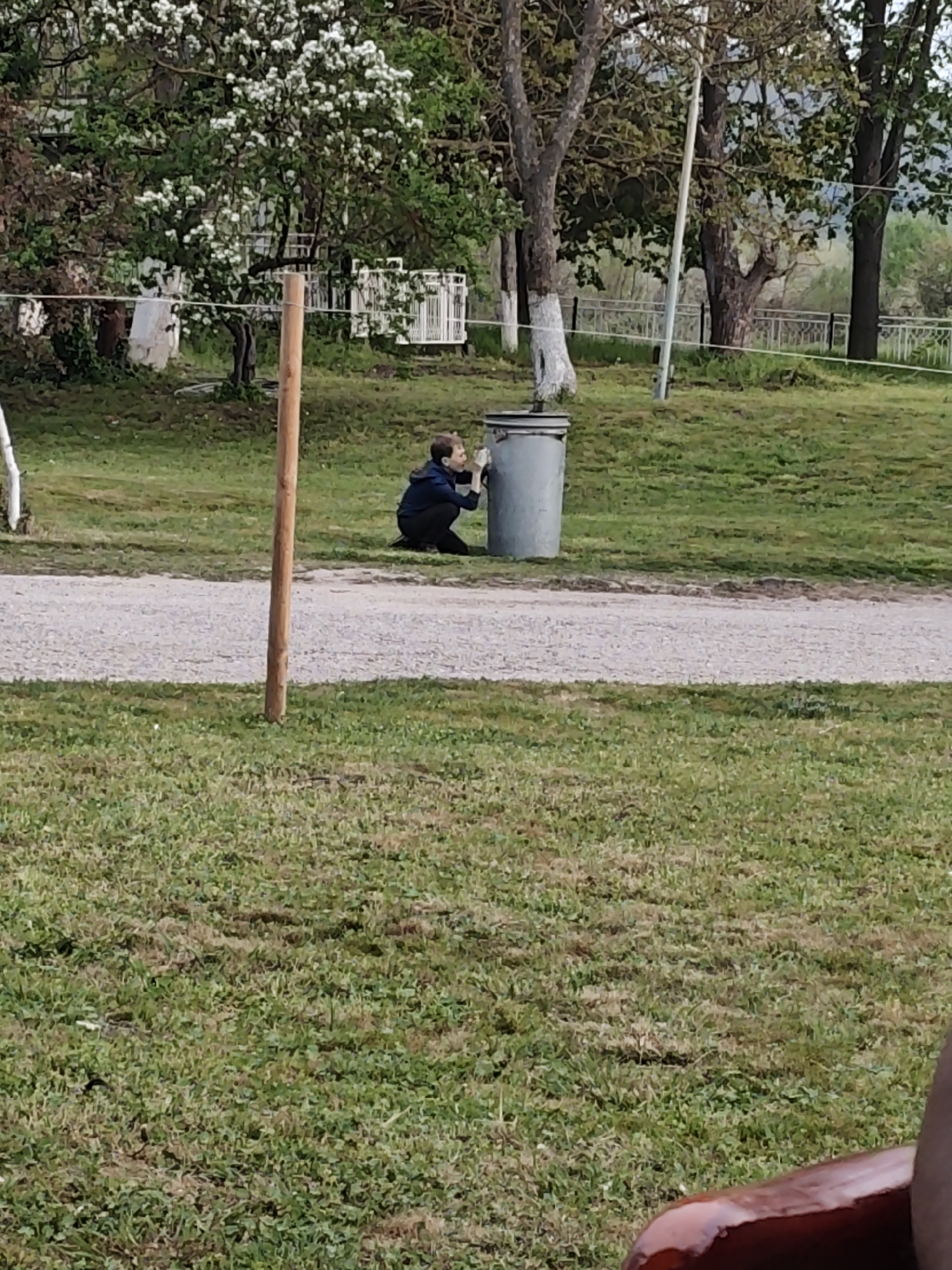 The image shows an outdoor scene in an area with trees and green grass. A young person is crouching next to a gray trash can. There is a dirt road running through the scene. A wooden post is visible near the road. In the background, there are trees with white flowers, a white fence, and more trees. The location appears to be a park or outdoor recreational area.