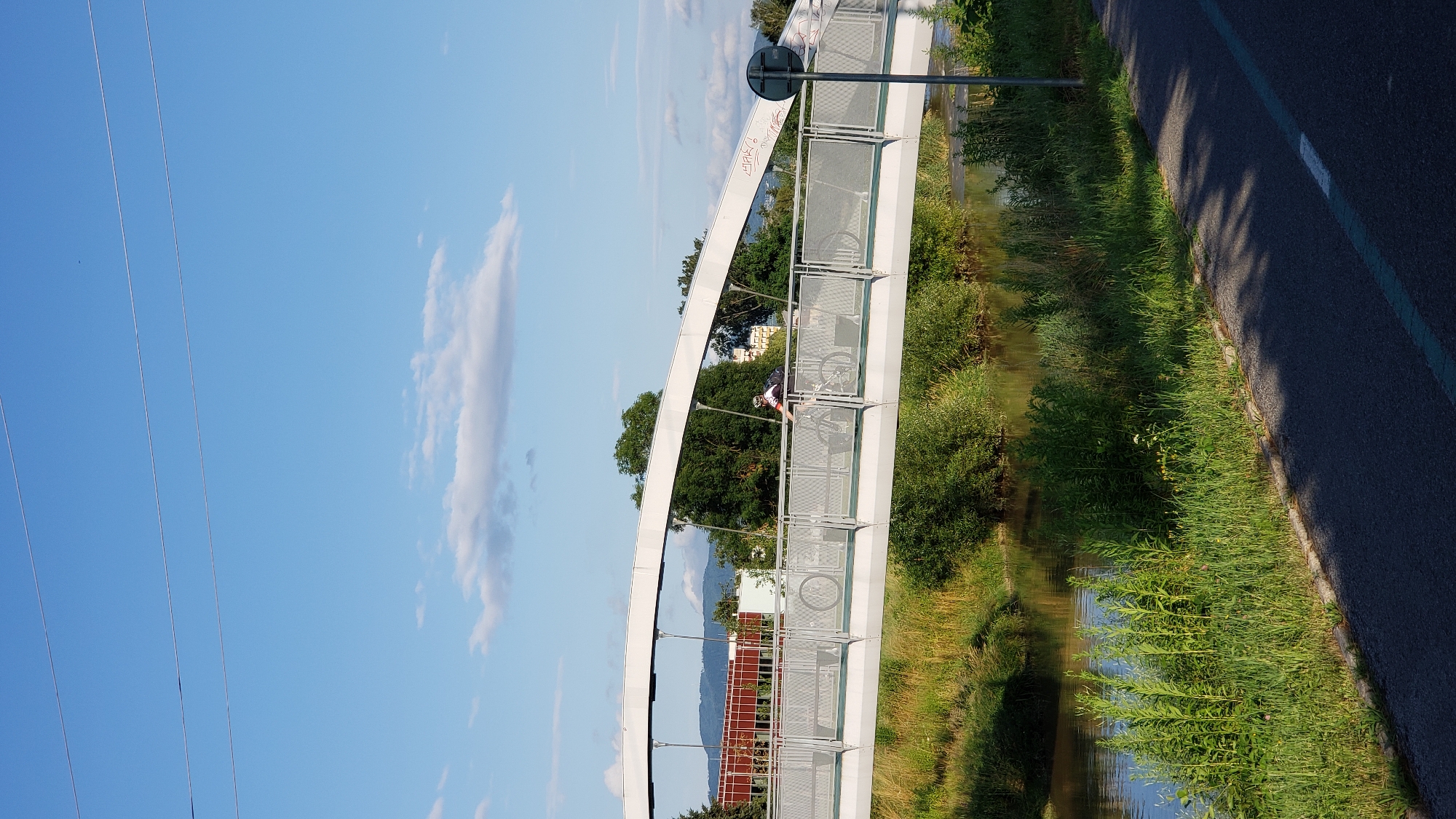 The image shows a pedestrian and cyclist bridge over a body of water, likely the Torysa River in Prešov, Slovakia. A cyclist is seen in motion on the bridge. The bridge is modern in design with a white arched structure and a metal railing. The surrounding environment is lush and green, with trees and vegetation on the riverbanks. A paved pathway runs alongside the river, casting shadows from the trees. The sky is blue with scattered clouds, and the overall scene suggests a pleasant day in an urban park or recreational area.