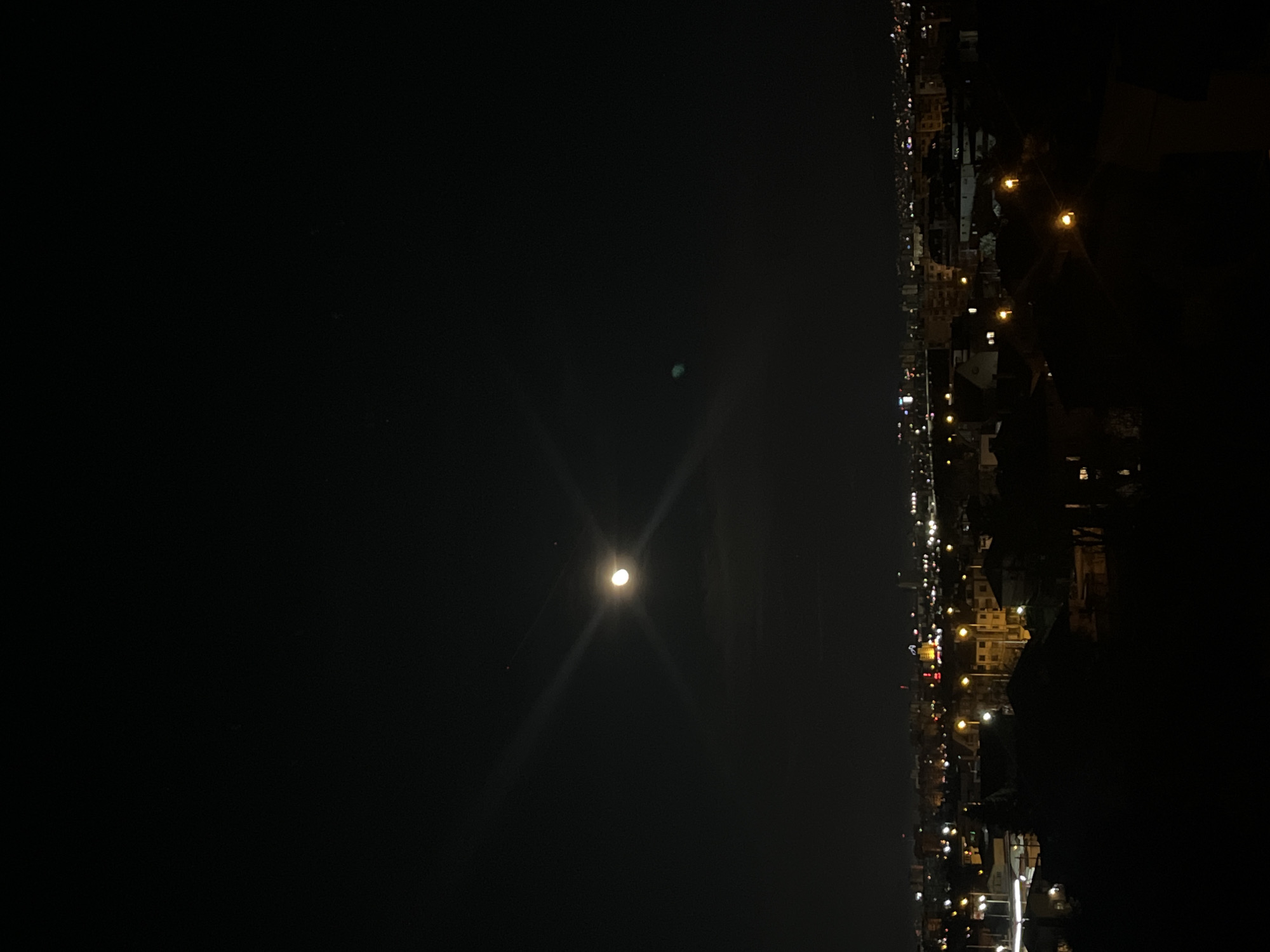 A low-angle, nighttime cityscape oriented vertically. To the right is the skyline of a city, defined by street lights and building lights, punctuated by dark shapes of buildings. To the left is the dark expanse of the night sky, in which is a bright, round moon with flares radiating from it. The environment is dark and suggests an urban setting at night. There are no people visible in the image. The city in the picture is NOT Sofia, Bulgaria.