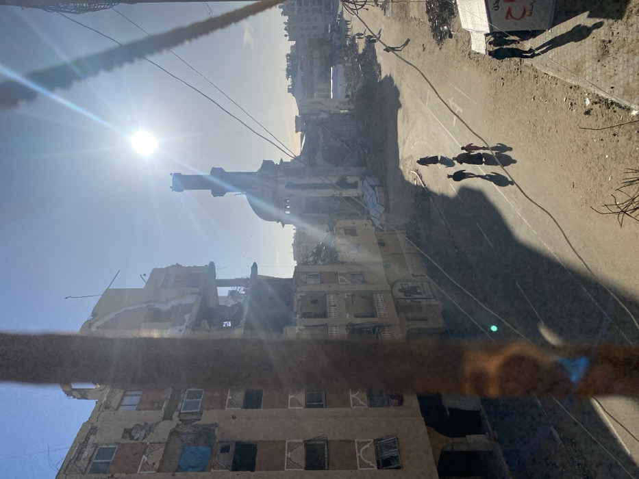 The image captures a scene of extensive urban devastation, likely taken from an elevated perspective overlooking a street in Khan Yunis, Palestinian Territory. The photograph is oriented 90 degrees clockwise; when viewed upright, a bright, intense sun is visible in the upper left of the clear sky, casting long, stark shadows towards the right. This suggests either early morning or late afternoon.
The scene is dominated by widespread destruction. Along a wide, possibly unpaved or debris-strewn street, buildings on both sides are in ruins. Many structures are partially collapsed, revealing exposed interiors, broken windows, and significant structural damage. Piles of rubble and debris are scattered across the area where buildings once stood. A prominent mosque, identifiable by its dome and a tall, slender minaret, stands notably damaged but largely intact towards the mid-left of the re-oriented frame.
On the street, a group of approximately five to six people, including one person wearing red, are walking on the right side, their elongated shadows stretching before them. Other figures or shadows are faintly visible further down the street. Their presence suggests ongoing human activity amidst the ruined environment.
In the immediate foreground, a thick, dark, horizontal bar or beam partially obscures the lower portion of the view, hinting at the vantage point of the photographer, possibly from a window or balcony. On a partially visible sign or billboard situated on the upper right side of the street, some Arabic text is discernible, including "الجمعية" (Al-Jami'yah), meaning "The Association" or "Society." The overall atmosphere is one of profound destruction and human resilience.