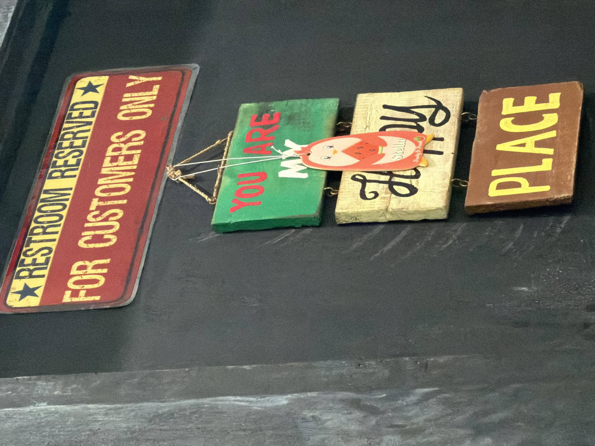 A metal sign with the words "RESTROOM RESERVED FOR CUSTOMERS ONLY" is hanging on a dark wall. Below it, there are three smaller wooden signs. The top one is green with red lettering that says "YOU ARE MY." The middle sign is light brown with black cursive writing that says "Happy." A small oval tag with a cartoon character and the word "Stella" is hanging from the middle sign. The bottom sign is brown with yellow lettering that says "PLACE." The overall scene appears to be a decorative display in a commercial establishment.