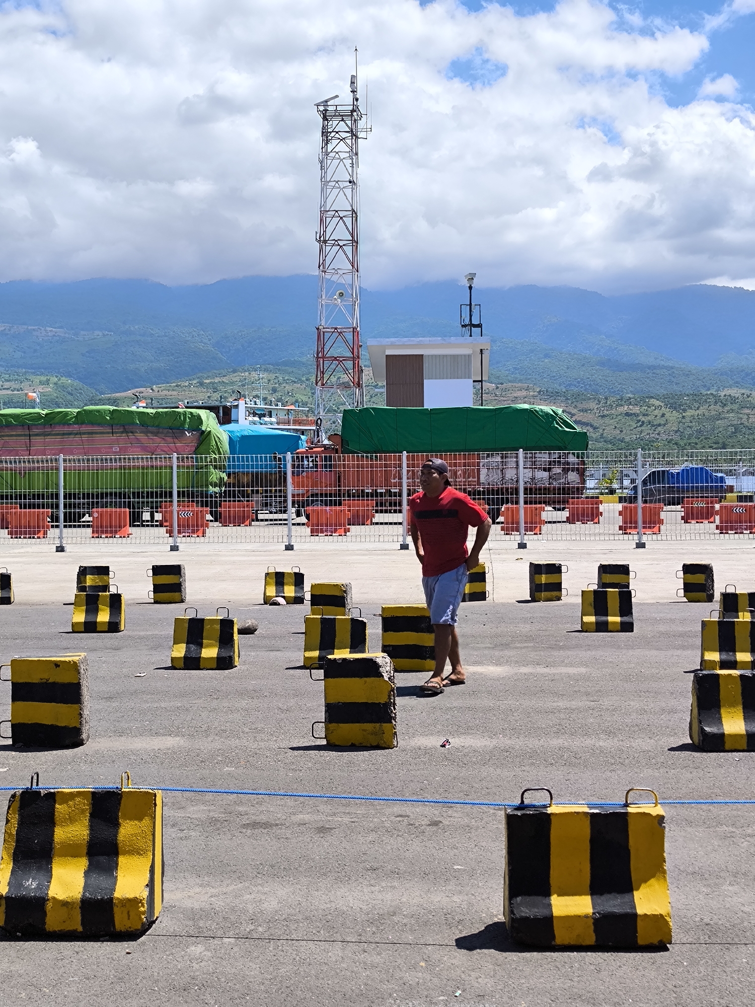 An eye-level, full shot shows a man standing in a paved lot among concrete barriers painted with black and yellow stripes, in the city of Gunung Malang, Indonesia. The man, centered in the image, wears a red shirt, light blue shorts, and brown sandals. He has his hands on his hips and appears to be looking to his left. The barriers are distributed unevenly across the lot, with a blue rope stretched between several of them in the foreground.
Behind the barriers, a chain-link fence separates the lot from an area with parked trucks, some of which are covered with green tarps. Behind the trucks, a red and white communications tower stands near a small building. A backdrop of mountains stretches across the horizon beneath a sky filled with puffy white clouds. The scene is well-lit, casting shadows from the barriers and the man. The environment suggests an industrial or commercial area, likely a port or transportation hub.