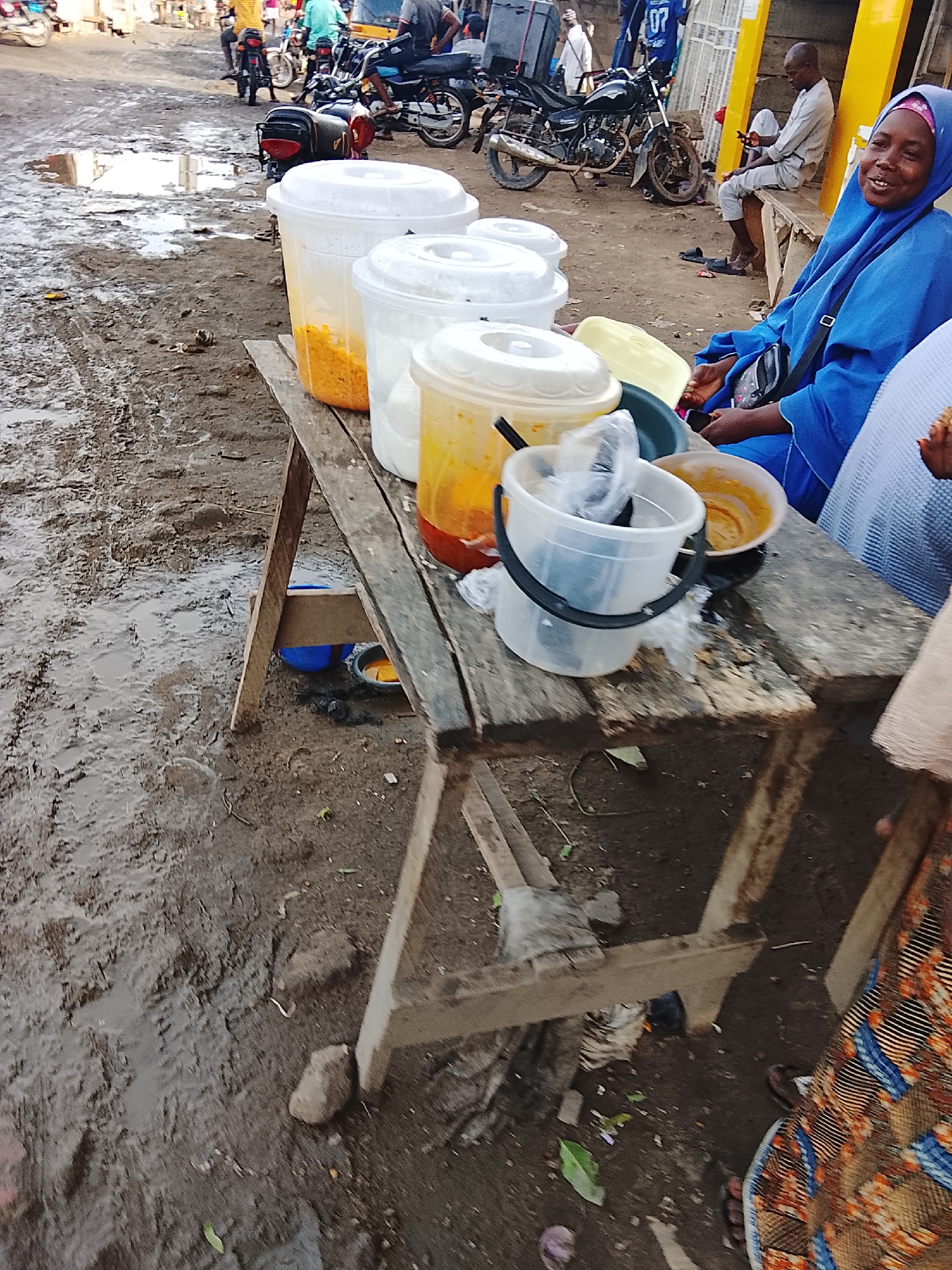 The image shows a food vendor and her setup on a muddy street in Abuja, Nigeria. The vendor, a woman wearing a blue hijab and matching tunic, sits behind a makeshift wooden table displaying several large transparent plastic containers filled with food items, some of which are yellow and orange. Other buckets and bowls contain additional ingredients. A customer, partially visible on the right, wears a colorful patterned fabric. In the background, there are parked motorcycles, and people are visible on a bench and walking in the distance. The ground is muddy with visible tire tracks and puddles. The light suggests it is daytime.