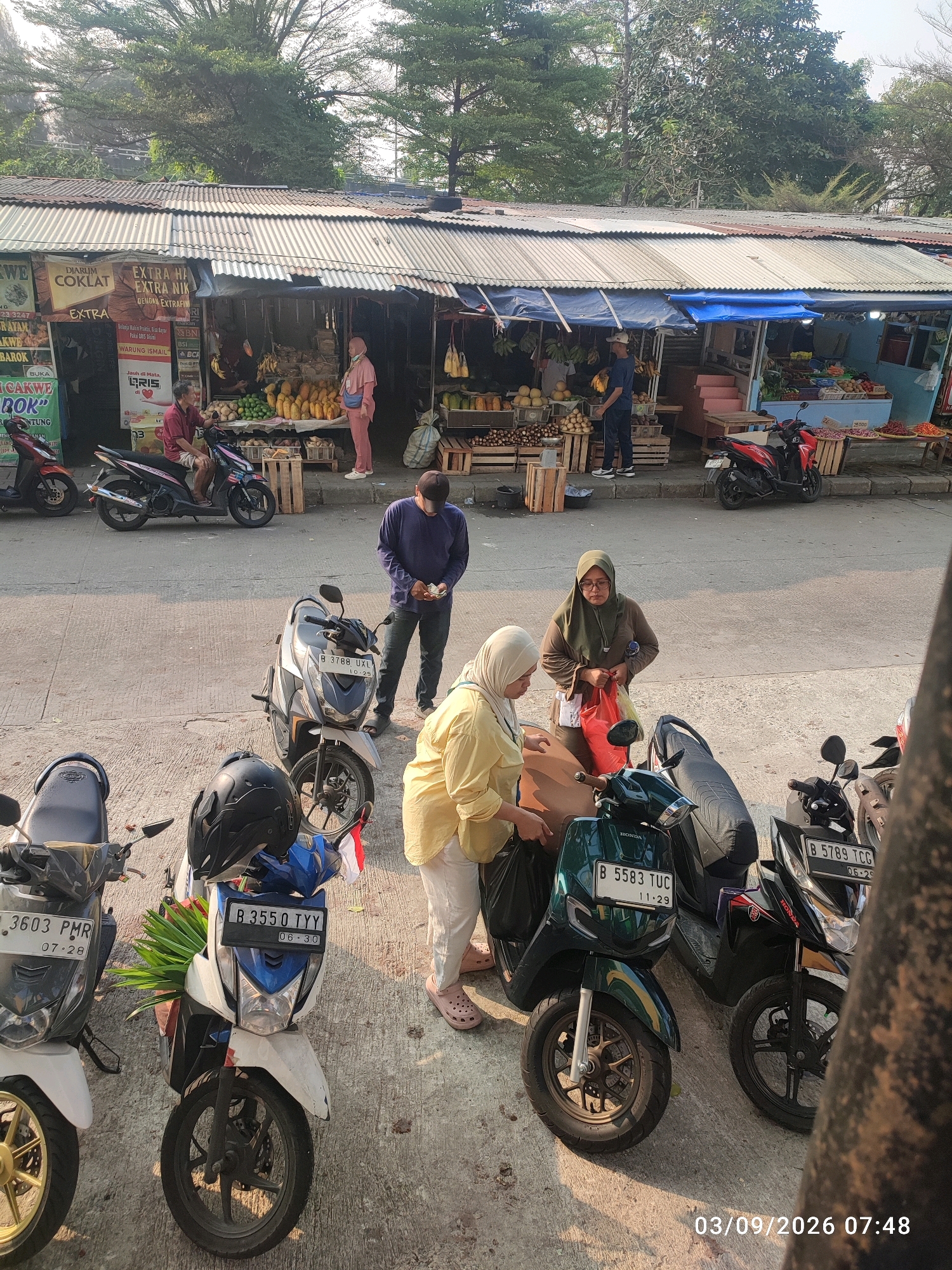 An outdoor street market scene in East Jakarta, Indonesia, is depicted in this image. The foreground features several parked motorcycles, some with helmets and bags. In the middle ground, two women are interacting with a motorcycle, one of whom is holding plastic bags. A man in a dark blue shirt and jeans is standing nearby, appearing to hold money. Further back, a row of market stalls with corrugated metal roofs is visible, displaying various fruits and vegetables. Several people are browsing or working at the stalls. Trees and foliage form a backdrop to the market. The ground is paved with concrete. The image is dated and time-stamped 03/09/2026 07:48.