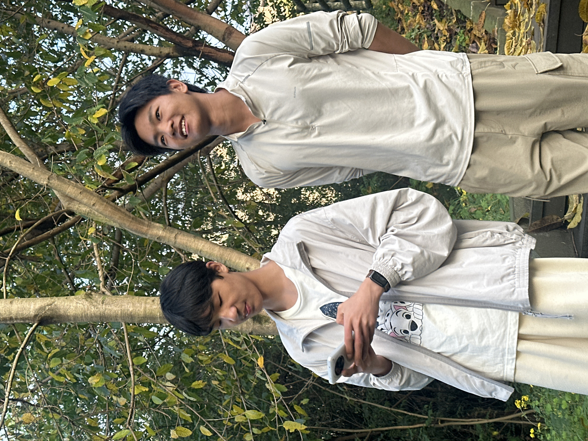 Two young men are outdoors surrounded by trees with green and yellow leaves, potentially in Jiangjin, China. The person in the background wears a light-colored, button-down shirt and matching cargo pants, smiling and looking slightly upward. The person in the foreground wears a Dalmatian-themed shirt with a jacket and light-colored pants, holding a smartphone in both hands. The background is a mix of branches and foliage.
