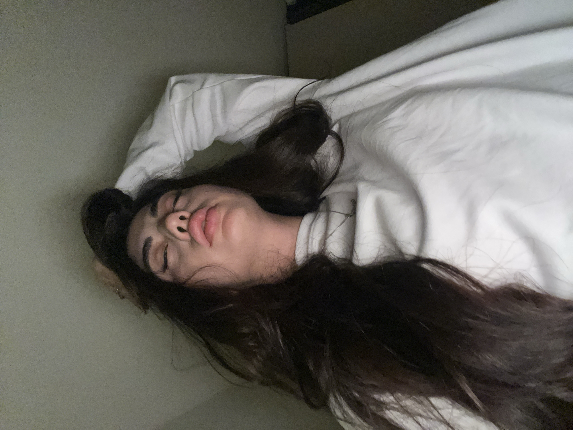 A low-angle, close-up shot shows a person lying down with their head tilted back and one arm raised, touching their hair. The subject has long, dark, wavy hair, and is wearing a white, long-sleeved shirt. Their eyes are partially closed, and their lips are slightly parted. The background is a plain, light-colored wall. The lighting is dim, casting shadows and creating a moody atmosphere. There are no discernible elements in the image that would suggest a specific location in the city of Chotěšov, Czechia.