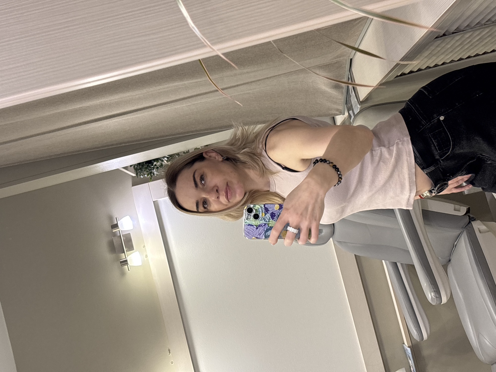 A woman is taking a selfie indoors. She has blonde hair and is wearing a light pink top and black pants. She is holding a phone with a floral patterned case. There is a dental chair visible in the background. The location is likely a dentist's office. The walls are painted in neutral tones, and there are light fixtures on the wall. The image does not contain enough information to determine if the location is in Vsevolozhsk, Russia.