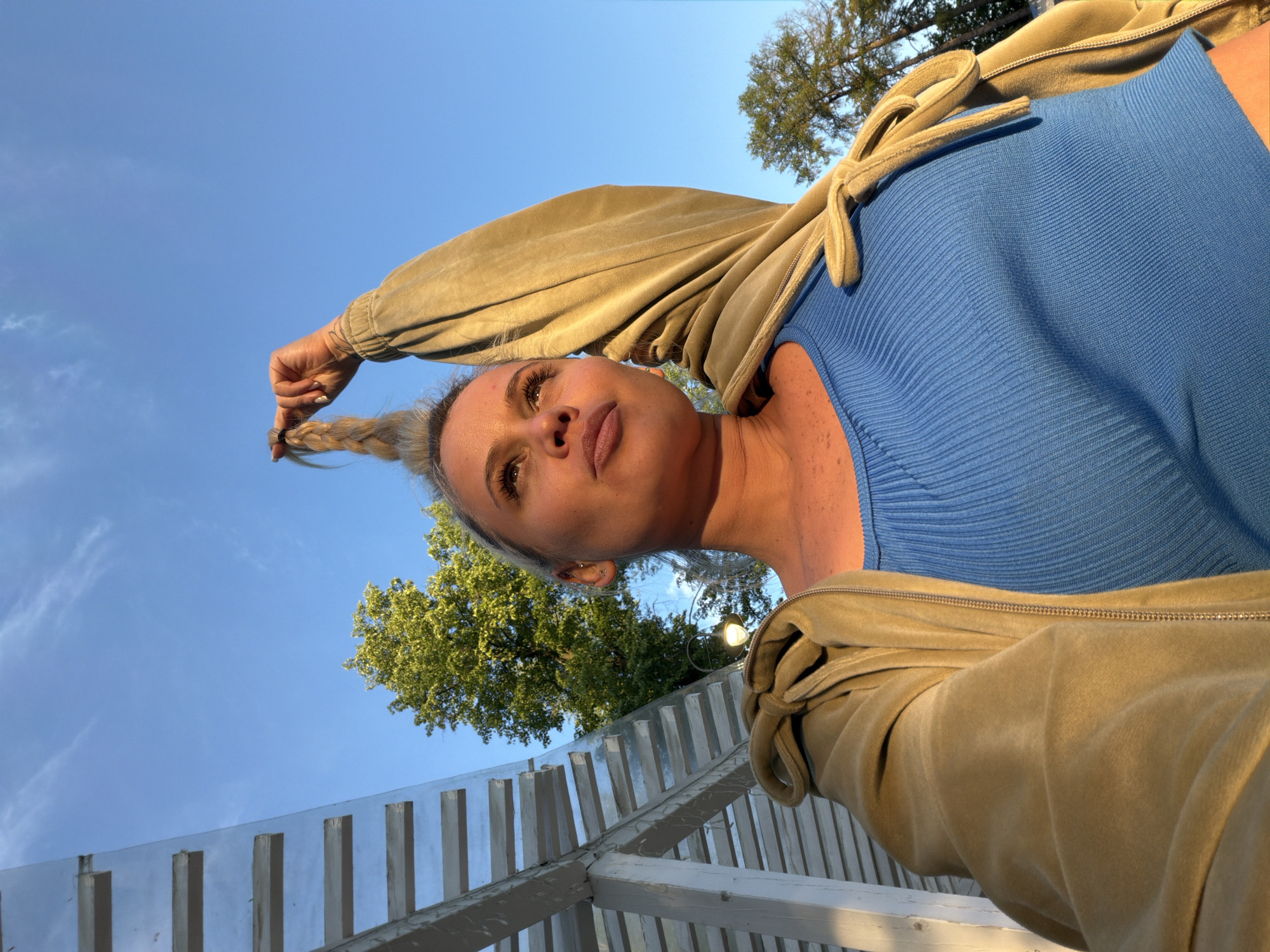 A low-angle, portrait-style shot captures a blonde woman in Sarapul, Russia. She stands outdoors against a blue sky partially obscured by a treetop. The woman's face is slightly angled to her left, and she holds the end of her braided ponytail. She wears a blue top and a tan zip-up jacket. Below her and the tree is a partially visible white wooden structure.