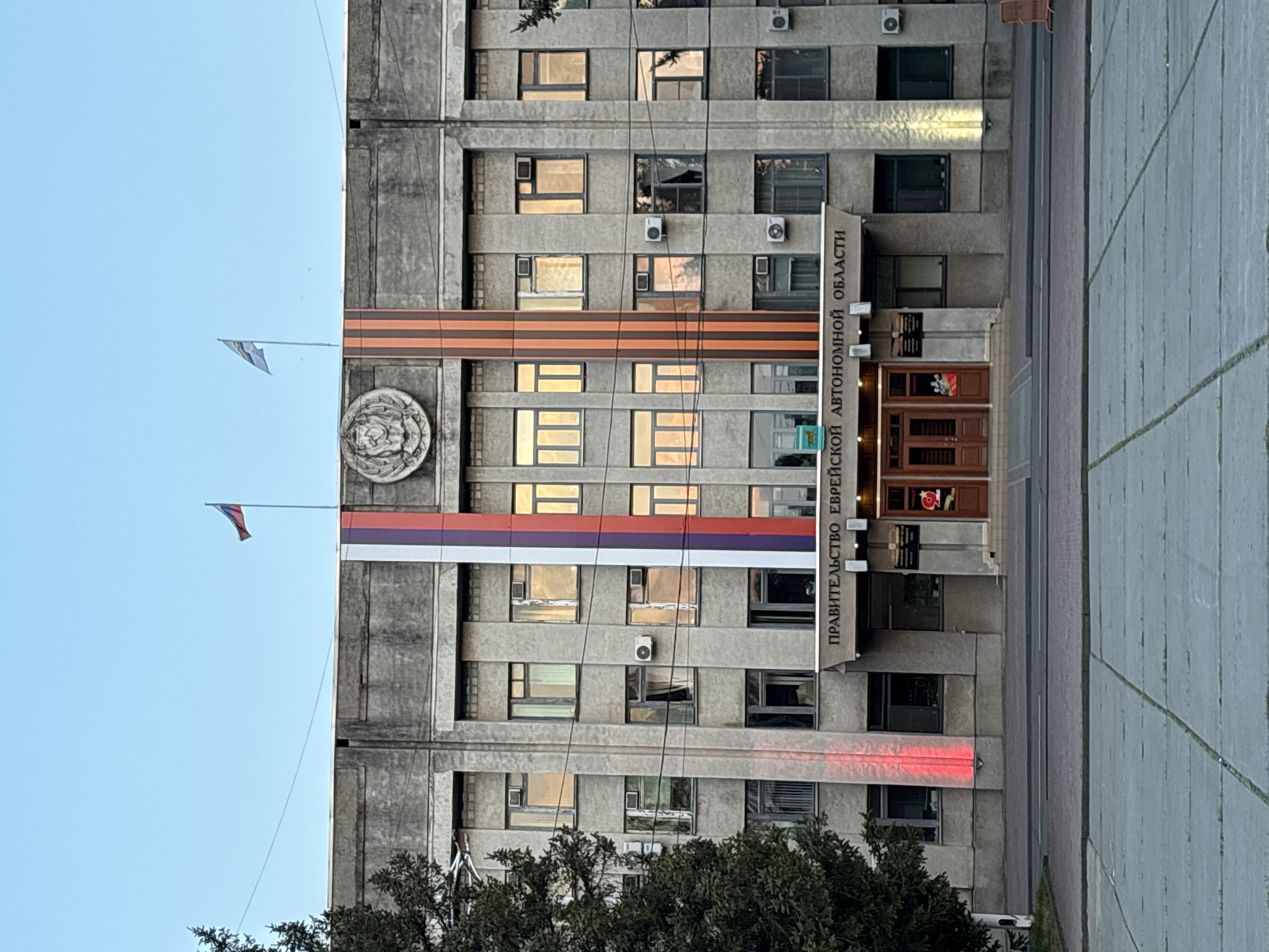 The image shows the building of the government of the Jewish Autonomous Oblast in Birobidzhan, Russia. The building is made of gray concrete and has multiple windows. The building is decorated with the flag of Russia and the Ribbon of Saint George. There are also the flags of the Jewish Autonomous Oblast. The building is located on a paved area. There are trees in front of the building.