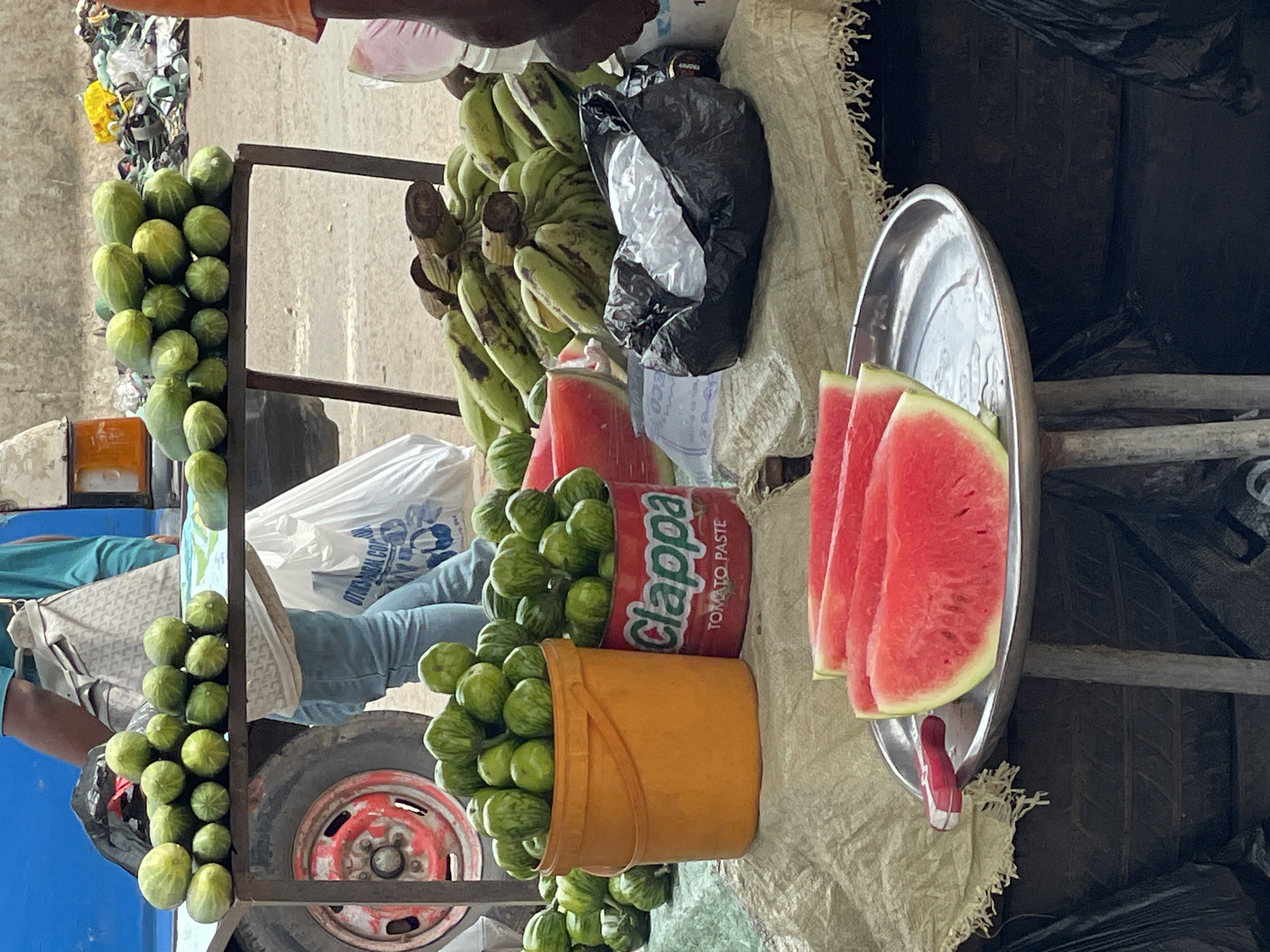 The image depicts a roadside market scene in Nkpor, Nigeria. A metal cart on wheels displays an array of produce for sale. Bunches of green bananas, watermelons cut into slices on a metal tray, and small, green, round eggplants are prominently featured. The eggplants are piled in an orange plastic bucket and stacked against a can of "clappa" tomato paste. A black plastic bag sits amongst the produce. A blue vehicle and a person wearing blue jeans can be seen in the background. The cart appears to be parked on a dirt or paved surface.