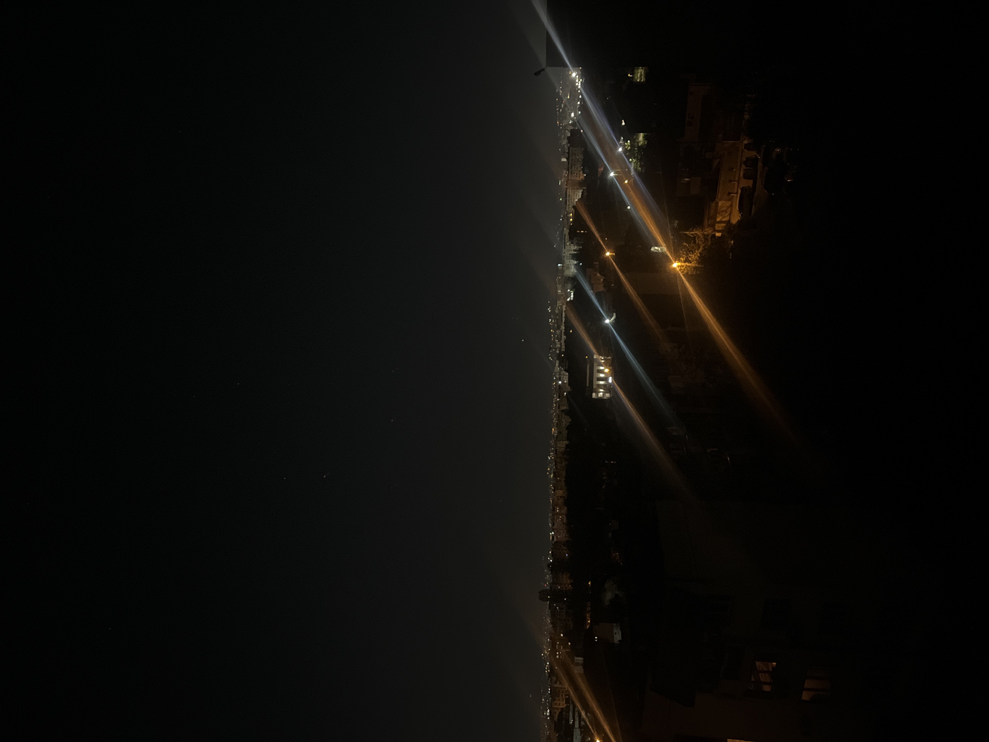 The media file displays a panoramic night-time view of a city, likely taken from an elevated position overlooking the urban landscape. The scene is dominated by a vast expanse of dark night sky in the upper portion, meeting a distant horizon line where city lights begin.
The lower half of the image showcases a sprawling urban area, identified by the context as Sofia, Bulgaria. Numerous artificial lights from buildings, streetlights, and infrastructure illuminate the city. These lights create distinct points and lines of varying colors, including bright white, cool blue, and warm orange/yellow. Many of the closer light sources exhibit prominent lens flares or streaks, radiating outwards and adding a dynamic visual effect to the night scene.
Silhouettes of buildings and structures are discernible amidst the illuminated urban fabric. The arrangement of lights suggests roads, streets, and clusters of buildings forming the city grid. There are no identifiable individuals or specific activities taking place; the image captures a static, wide-angle view of the city asleep.
The time of day is clearly night, characterized by the dark sky and the reliance on artificial illumination. The weather appears clear, allowing for a broad, unobstructed view of the distant city lights. No visible text or specific landmarks are clearly identifiable within the image itself to confirm the location, but the context indicates it is Sofia. The overall mood is one of quiet grandeur, showcasing the expansive scale of the urban environment at night.