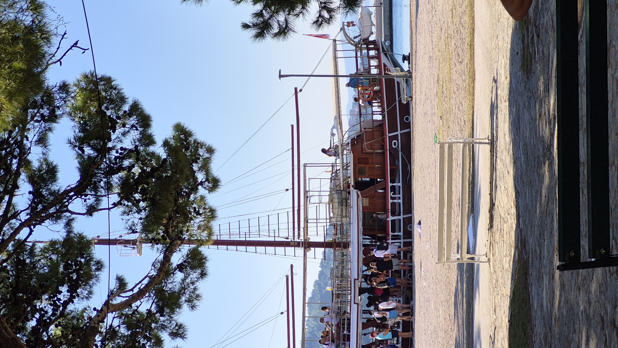 A wooden sailboat is docked at a pier with people boarding. The boat has multiple decks with railings and tables. A few people are standing on the beach in front of the boat, while others are waiting to board. The scene is set on a sunny day with a clear blue sky. In the foreground, there is a white bench and a dark green bench. The background shows a body of water and a tree with pine cones.
