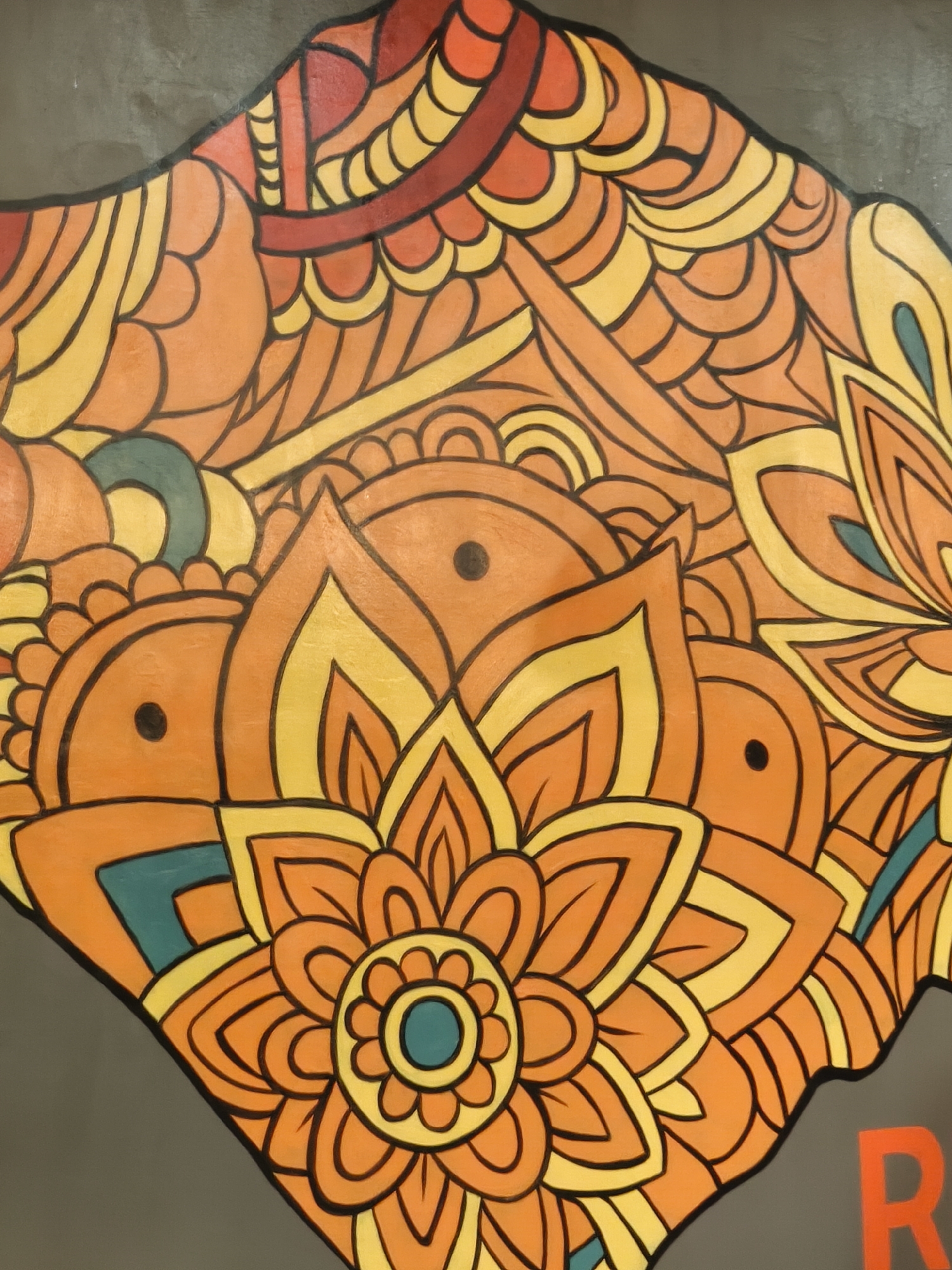 This is a close-up, overhead shot of a painting or mural featuring a vibrant, symmetrical, floral-inspired design in shades of orange, yellow, and teal against a dark gray background. The artwork is abstract and ornamental, with intricate patterns resembling petals and swirling lines. There are no people or discernible objects other than the painted design. The style is reminiscent of a mandala or a detailed folk art pattern. The visible elements suggest a decorative or artistic installation. A single, bright orange letter "R" is visible on the far right edge of the image, partially cropped. No specific time of day, weather, or emotional cues are evident. The location context of Kuta, Indonesia, does not provide specific visual cues within the image itself, but suggests a possible public art piece in that region.