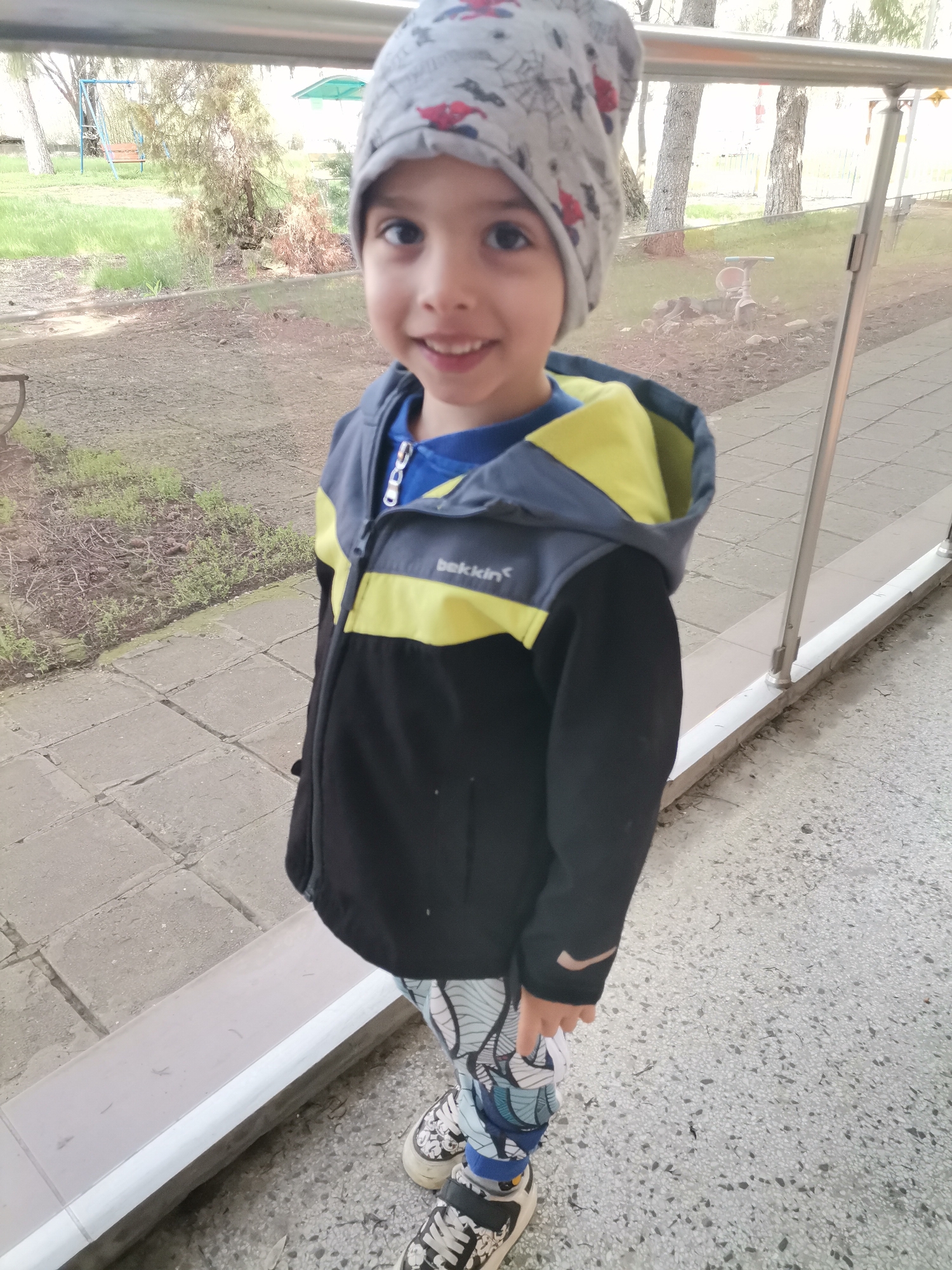 A young boy stands outdoors in front of a railing. He wears a patterned gray beanie, a blue shirt, a black jacket with yellow accents, patterned pants, and black and white sneakers. Behind him are trees and a paved area. The image was likely taken in Dobrich, Bulgaria.