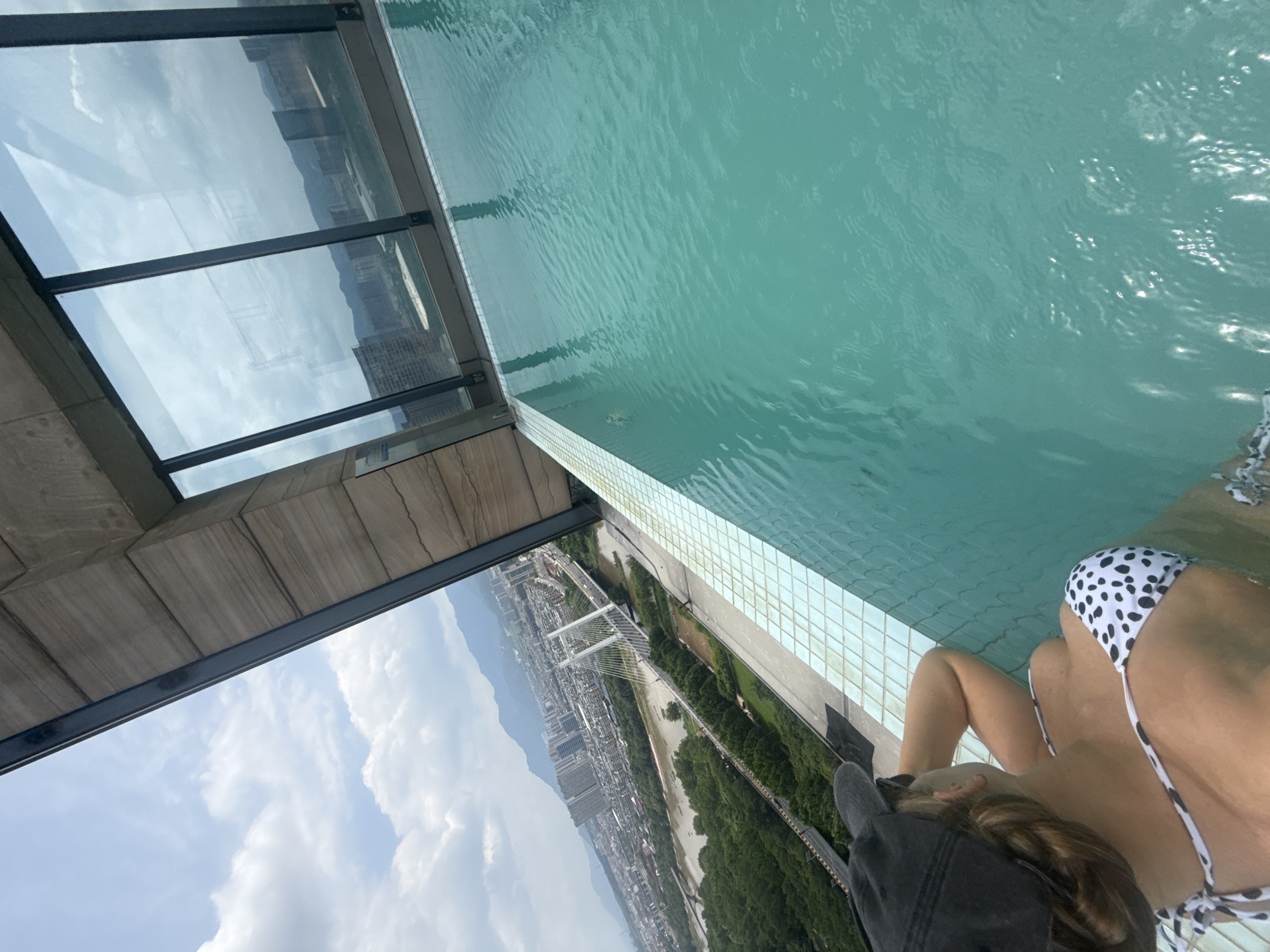 In the city of Yiwu, China, a person wearing a black baseball cap and a black and white polka dot bikini is seated in an infinity pool on a rooftop, overlooking a cityscape. The pool's turquoise water reflects the cloudy sky and the surrounding buildings. In the distance, a river with a modern cable-stayed bridge crosses it, flanked by lush green trees and urban development, with mountains forming the backdrop under a hazy sky. The pool edge is lined with light blue square tiles. The building's angled ceiling, made of wood-paneled material and metal beams, is visible in the upper right portion of the image, reflecting the sky in its large glass windows.