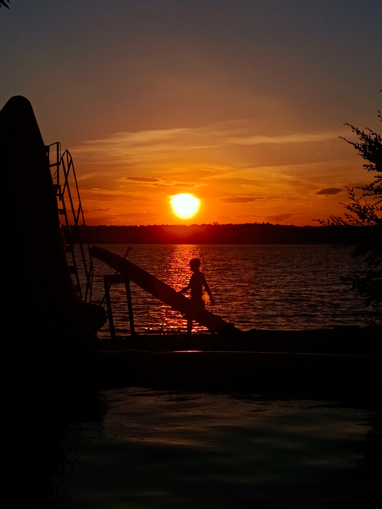 A silhouette of a person stands on a wooden slide next to a body of water during sunset. The sun is setting on the horizon, casting a warm orange and yellow glow across the sky and water. The person is facing the water, and appears to be either preparing to slide or has just emerged from the water. The scene is dark with the exception of the sun and its reflection on the water, creating a dramatic contrast. The overall mood is serene and peaceful, evoking a sense of summer vacation or a moment of quiet contemplation.