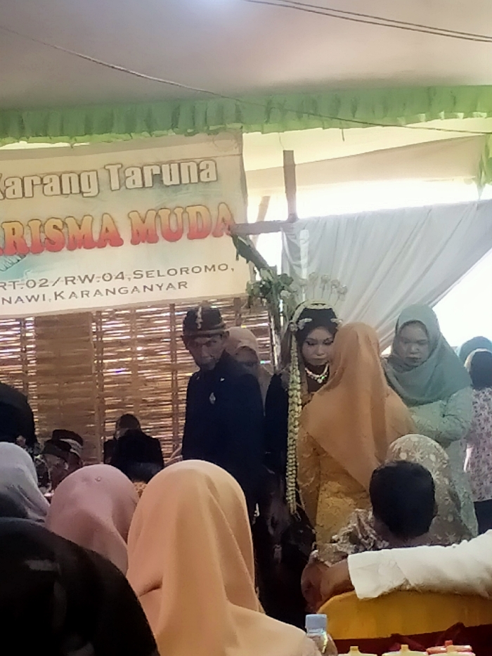 A wedding is underway, with the bride and groom standing in the center. The bride wears traditional attire with floral adornments in her hair, while the groom dons a formal dark suit and headwear. Attendees are seated, their heads and shoulders visible in the foreground, some wearing hijabs. Behind the couple, a banner reads "Karang Taruna RISMA MUDA RT-02/RW:04, SELOROMO, NAWI KARANGANYAR," suggesting the event is linked to a youth organization in the Seloromo area. The setting appears to be an open-air venue, indicated by the tent-like structure and natural lighting.