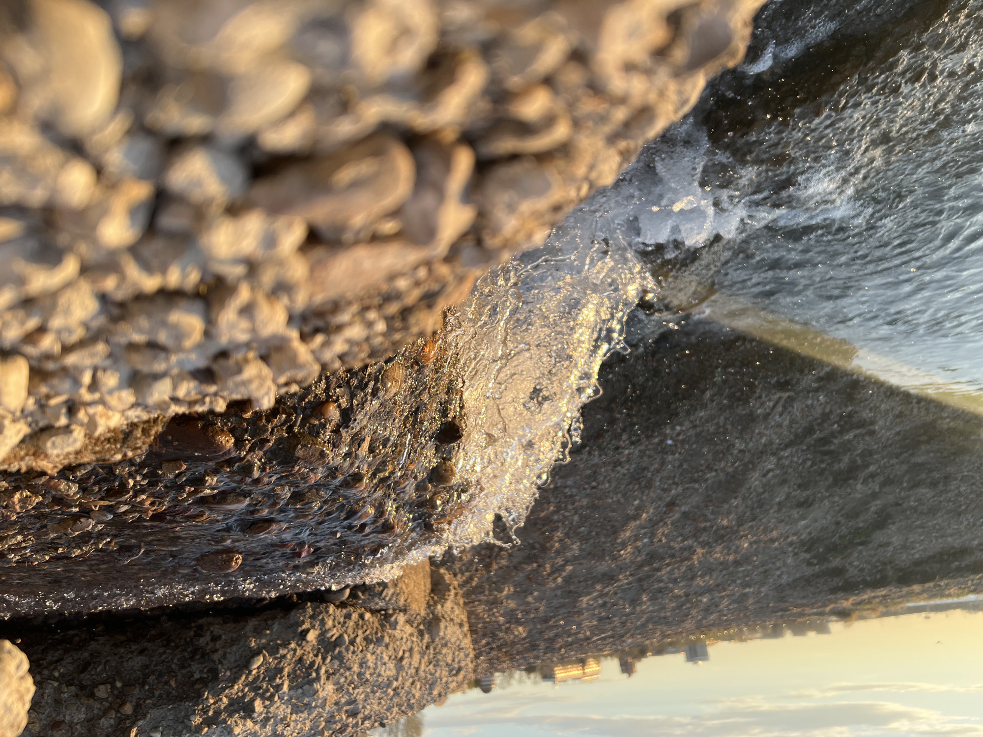 The image shows a close-up view of a textured, concrete structure, possibly a pier or seawall, with water splashing against it. The structure appears to be made of aggregate concrete, with pebbles and stones visible in the mix. The water is partially frozen, with ice formations apparent on the surface and edges. In the background, indistinct shapes suggest buildings and the sky, hinting at a cityscape. The light suggests it is either sunrise or sunset. Given the prompt's mention of Irkutsk, Russia, it is possible that this scene depicts the Angara River or Lake Baikal shoreline within the city. Specific objects such as people or specific buildings are not visible. The action is the water interacting with the concrete structure, partly freezing.