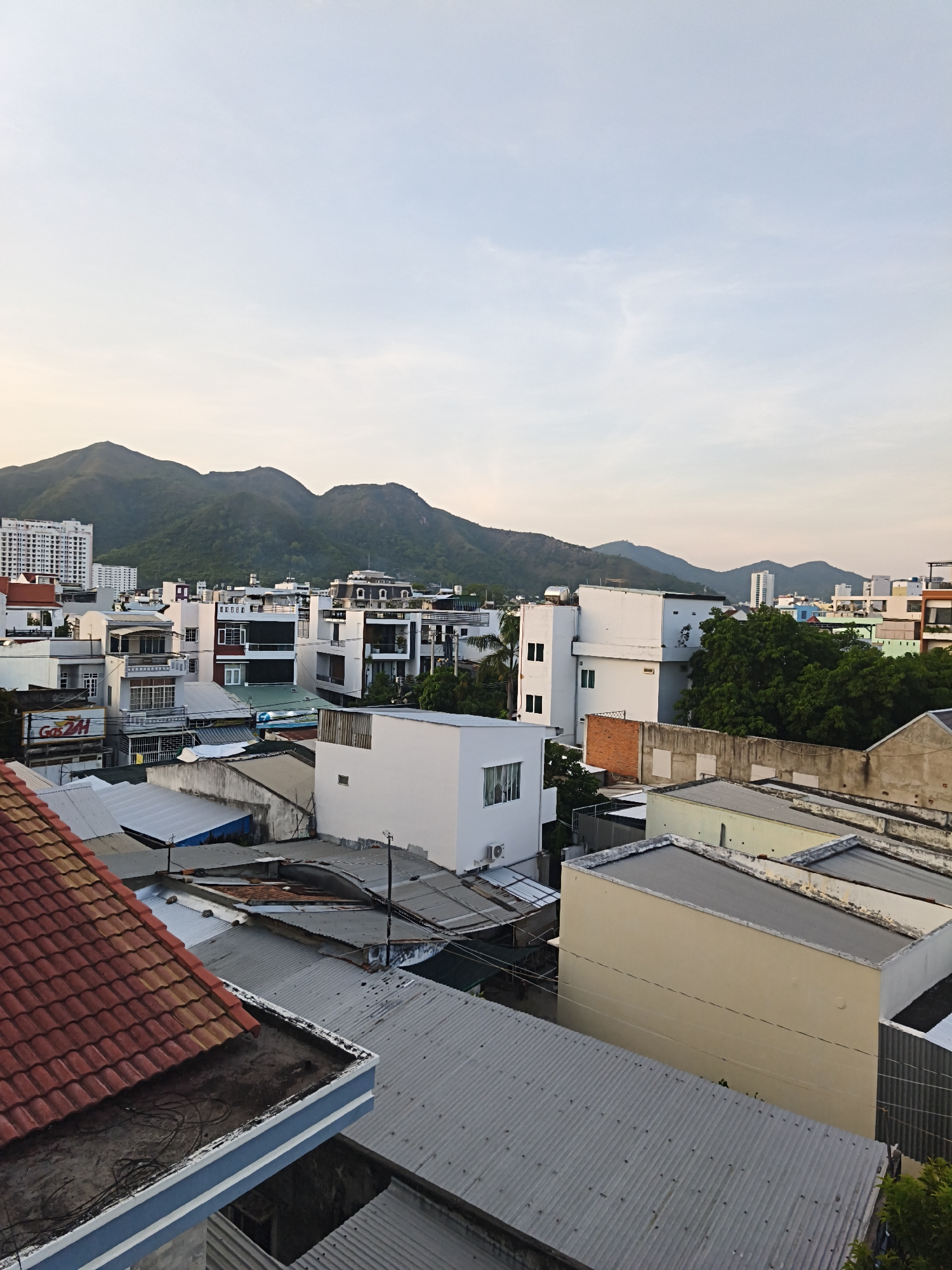 The image is an aerial view of a densely populated urban area, likely Bac Nha Trang Ward, Vietnam, at dusk. The foreground is dominated by the rooftops of various buildings. In the lower left, a red-tiled roof slopes downwards, revealing a dark, flat surface with visible wiring and a blue and white striped edge. Adjacent to this is a grey, corrugated metal roof. Further into the midground and background, a multitude of buildings of varying heights and architectural styles are visible. Many are painted white, some with black accents and balconies. There are also buildings with colorful facades, including light yellow and green. Several trees, predominantly green and lush, are interspersed among the structures, particularly in the midground.
In the background, a range of green, rolling mountains form a natural backdrop to the city. The sky above is a pale blue, transitioning to a warmer, lighter hue near the horizon, indicative of sunset or sunrise. The lighting suggests it is either early morning or late afternoon. There are no people visible in the image. The scene depicts a typical residential and commercial cityscape, characterized by its tightly packed buildings and proximity to natural landscapes.