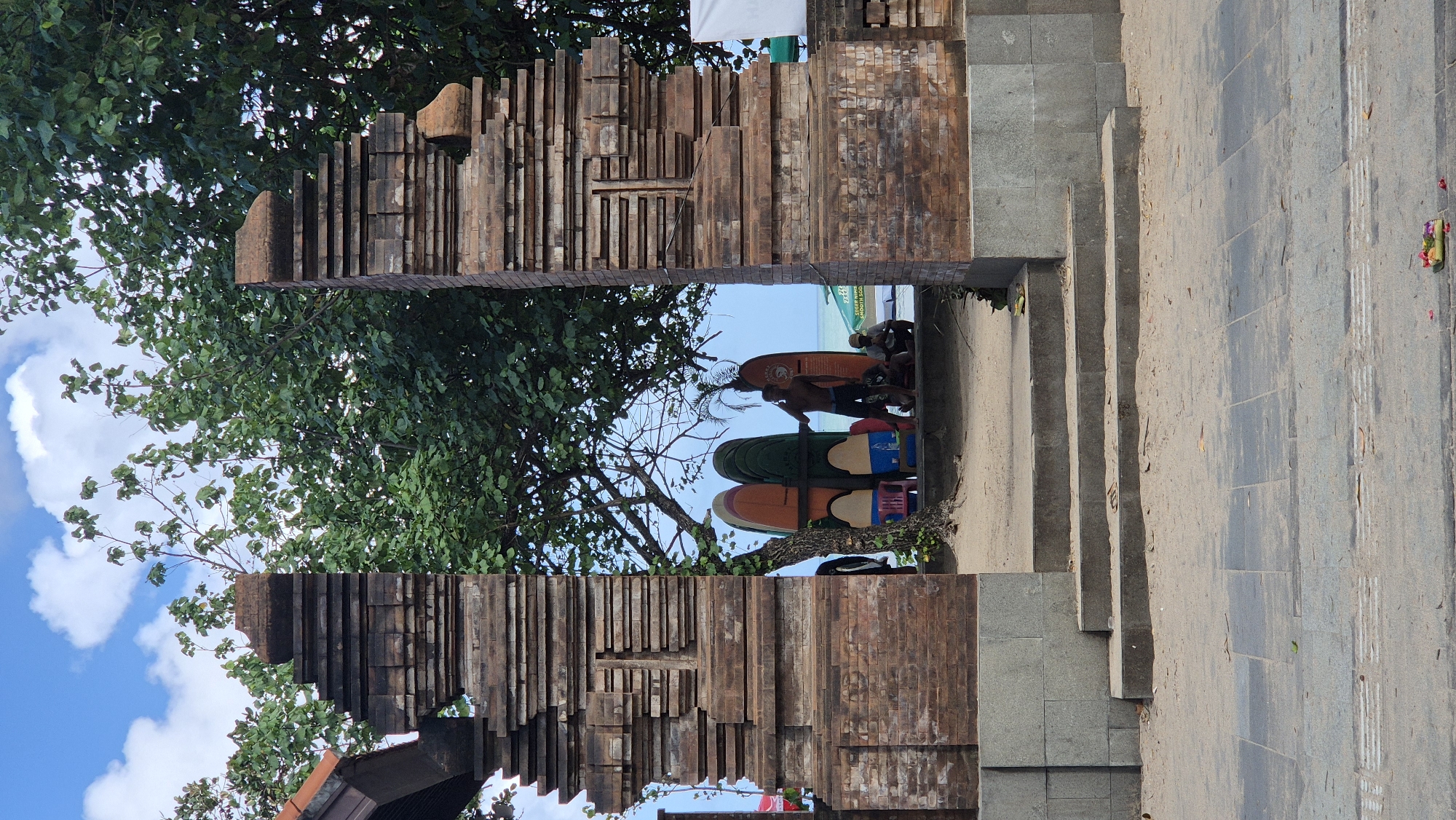 The image shows a view through an ornate wooden structure, likely a decorative entrance or archway, in Kuta, Indonesia. The structure is composed of multiple layers of stacked wooden bricks, with a gap in the middle that frames a scene on the other side.
Through the gap, one can see several surfboards propped up against a tree, with individuals gathered around them. The surfboards vary in color and size, with some being orange, green, and blue.
To the right of the wooden structure, there are concrete steps leading down, likely to a beach or lower area. The ground is paved with gray stone tiles.
The surrounding environment includes trees with green leaves, a blue sky with white clouds, and a glimpse of the beach in the distance. A traditional offering is present at the base of the stairway.