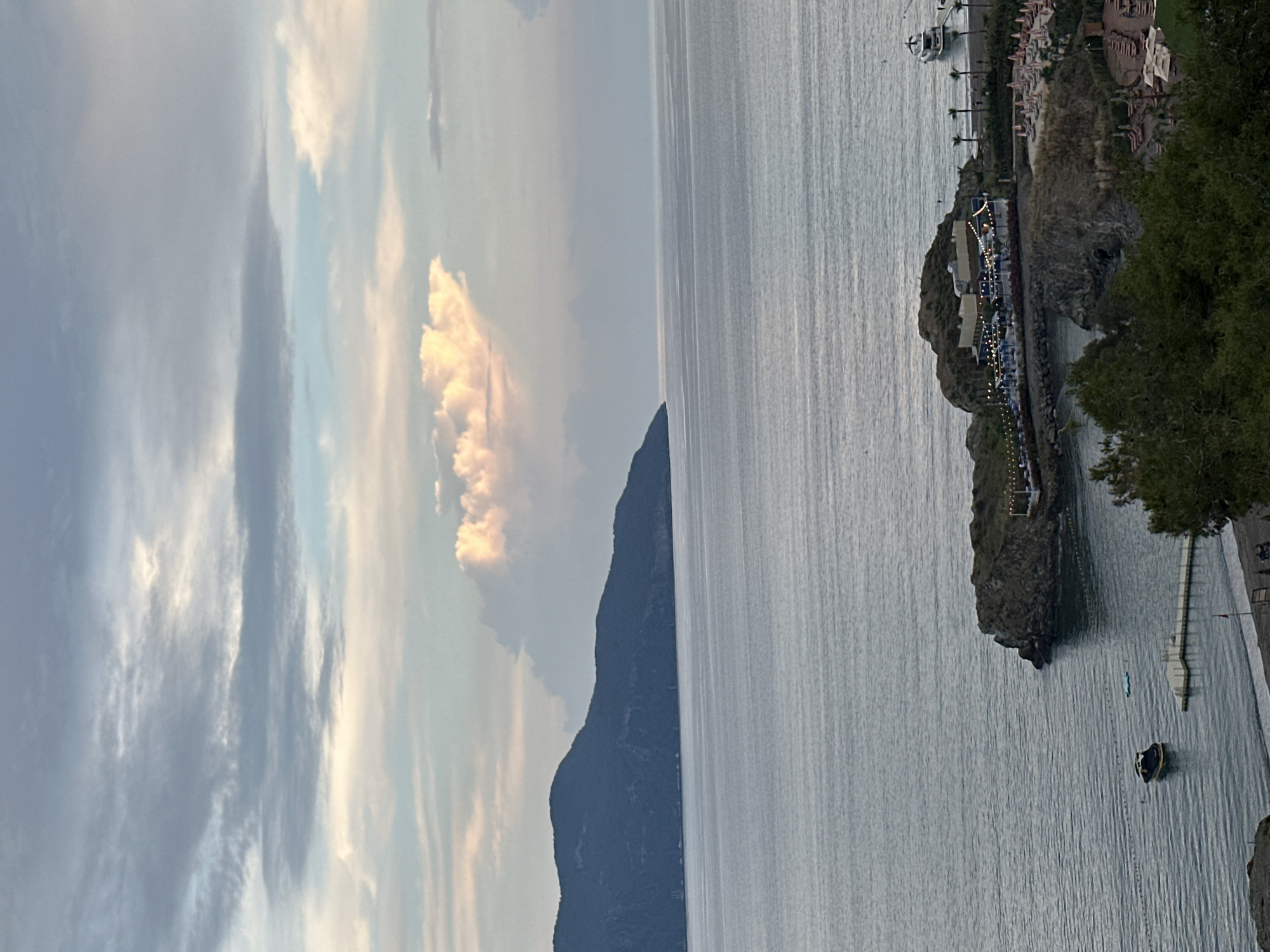 The image depicts a landscape with the sea on the right and the sky on the left. In the background, there is a mountain range, and a few white clouds float in the sky. On the sea, there is a small island with buildings. It is a coast with hotels in Fethiye, Turkey. A small boat is floating on the water.