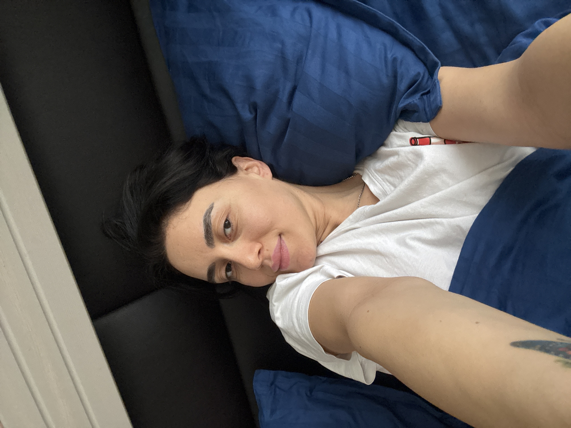 An adult female is depicted in a selfie, lying in a bed within what appears to be a bedroom. She has a medium skin tone, dark brown hair, and dark eyebrows, looking directly at the camera with a slight smile. She is wearing a white short-sleeved t-shirt and a silver-colored chain necklace. A tattoo, primarily dark blue/black with hints of red and green, is visible on her upper left arm. Her left arm is extended towards the camera, holding the photographic device. She is covered from the waist down by dark blue bedding, which also includes a pillow supporting her head. Behind her head is a black, padded headboard. Above the headboard, a white structure with horizontal slats is visible. The specific location in the city of Tbilisi, Georgia, cannot be determined from the visual information provided in the image.