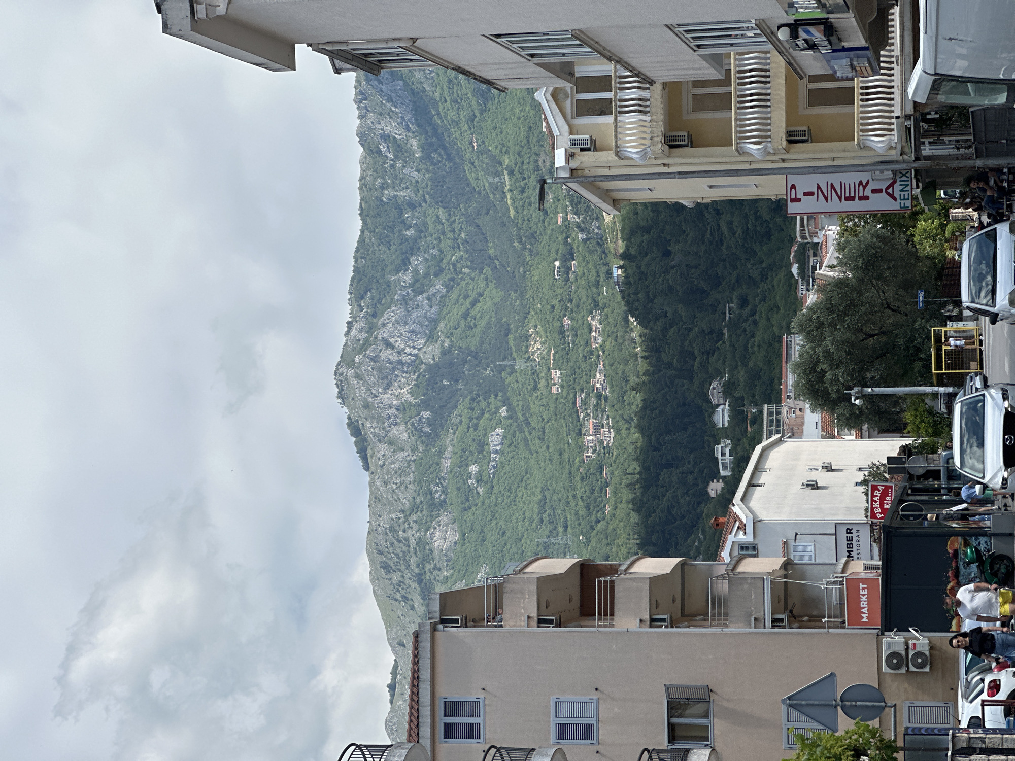 The image shows a street scene in Budva, Montenegro, looking toward a large mountain. Buildings line the street, and several signs are visible, including "PIZZERIA," "MARKET," and "MBER RESTORAN." People are walking along the street, and there are parked vehicles on the right side of the frame. The sky is overcast. The mountain dominates the left side of the image.