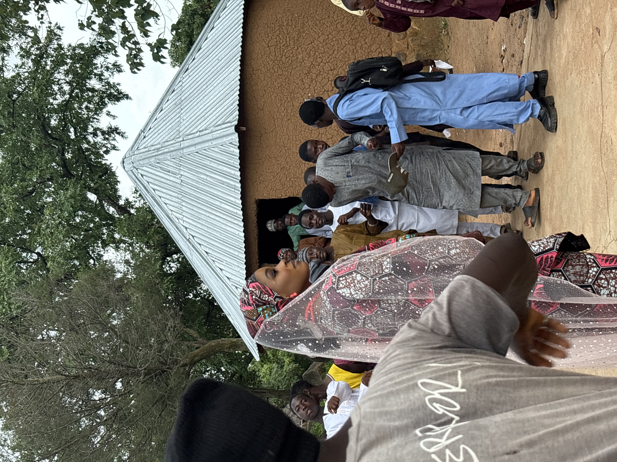 A group of people are gathered outside a mud-walled building with a corrugated metal roof. A woman in the foreground wears a colorful headscarf and a veil adorned with small reflective sequins. She is looking to her right. Other individuals, mostly men and boys, are dressed in traditional African attire and standing or walking around. Some are looking at the woman, while others are engaged in conversation or looking elsewhere. The ground is dusty and uneven. The building appears to be a simple dwelling or community structure. The overall scene suggests a social gathering or event taking place in a rural or semi-urban setting, possibly within the city of Jos, Nigeria.