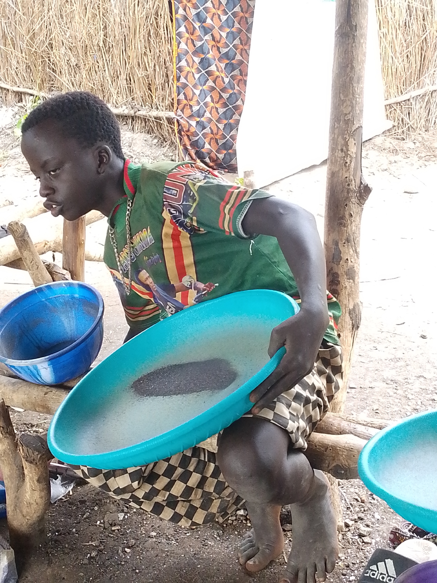 This image depicts a person sitting on a wooden structure, holding a large, turquoise-colored pan filled with dark material and water. The person is wearing a green shirt with colorful patterns and a chain necklace. Their lower body is covered by checkered shorts. To the left, a blue plastic bowl is visible, and to the right, another turquoise pan with white liquid is partially visible.
The background shows a thatched wall and a wooden pole. The ground is dirt and gravel. The scene appears to be outdoors, possibly in a rural or informal setting. The lighting suggests it could be daytime, with natural light illuminating the scene. The person's facial expression is neutral or slightly focused on the task at hand. The overall impression is one of everyday activity, possibly related to washing or processing materials. There is no visible text in the image.
