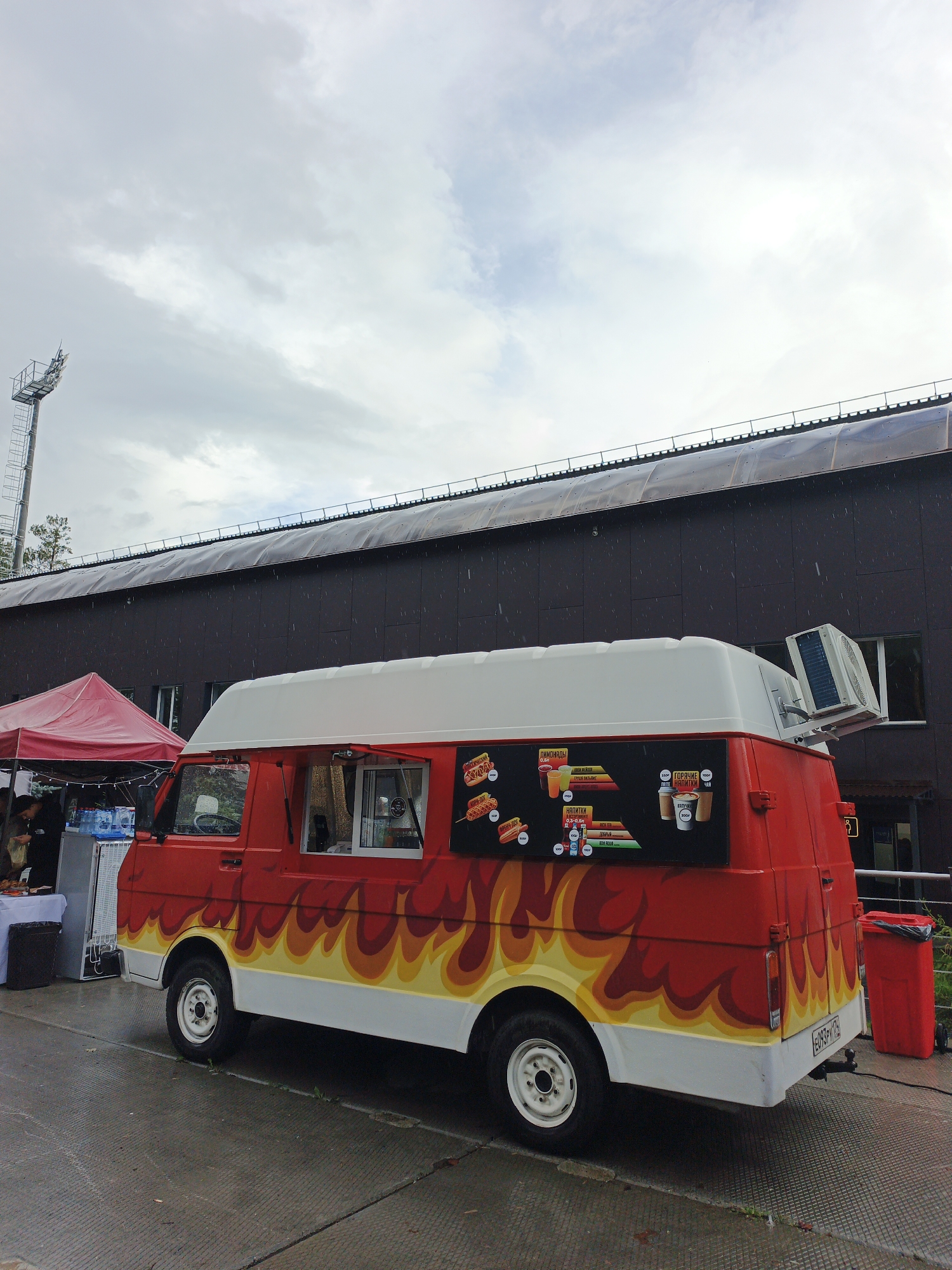 The image depicts a red and white food truck with a flame decal design parked outdoors. The food truck has a white roof with an air conditioning unit mounted on top. A menu with images of food and drinks is visible on the side of the truck. To the left of the food truck, a red tent is set up with tables and bottles of water. A tall light tower is visible in the background. The ground appears wet, suggesting recent rain. The scene is set against a large, dark building with a corrugated roof. The image was taken in Zlatoust, Russia.