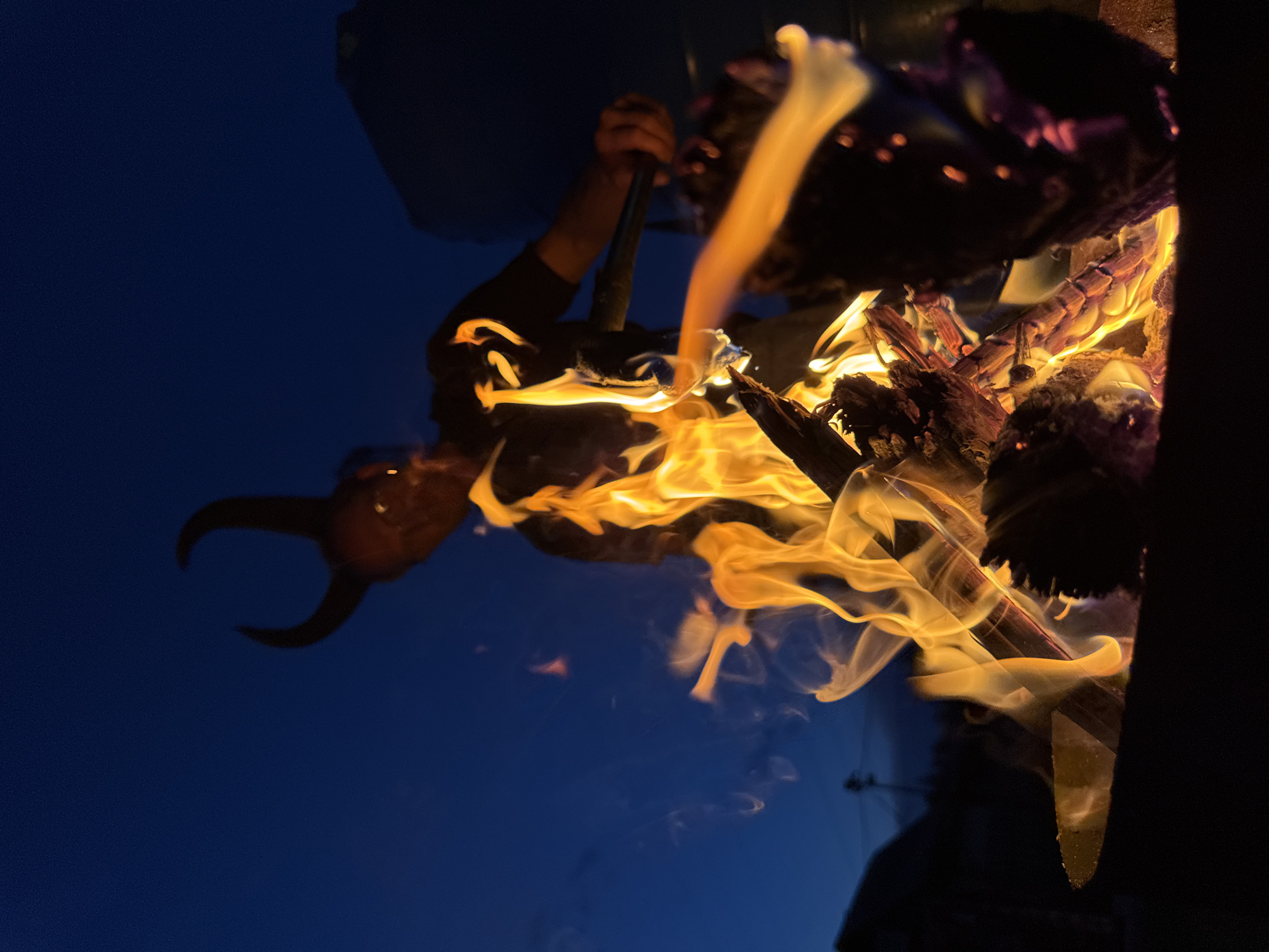 The media file shows a close-up, low-angle shot of a campfire at dusk, with a clear, dark blue sky as the background. The fire is actively burning, with bright orange and yellow flames licking at dark logs and branches. Smoke is also visible, rising and dissipating into the air.
Reflected in the smooth, dark surface below the fire, which appears to be a reflective material, is a distorted image of a person wearing a mask with horns. The mask is dark brown and has two prominent, curved horns. The reflection also shows a dark sleeve and a hand holding a stick or poker, suggesting the person is tending to the fire. The reflection is partially obscured by the flames and smoke.
The overall impression is one of mystery and a touch of the uncanny, due to the demonic-like mask reflected in the surface. The time of day is clearly evening or night, as indicated by the dark sky and the bright, active fire. The location context of Chaikovsky, Russia, does not provide specific visual cues within the image itself.