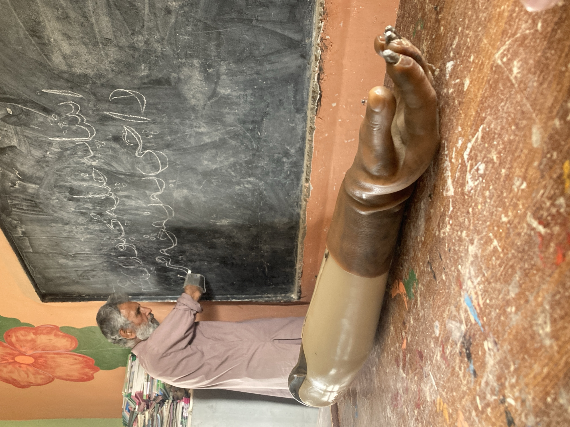 A classroom in an unknown city, unknown country, shows a man with gray hair and beard writing on a blackboard with white chalk. He wears a light brown shirt. On the wall is a drawing of a large orange flower. The image is cropped to the right with a prosthetic arm and hand.