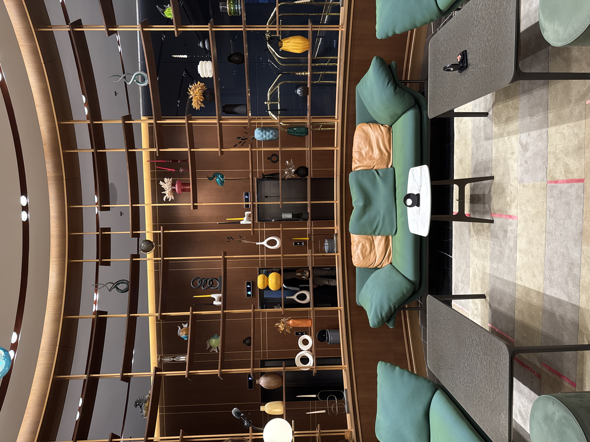The image shows an interior space featuring a tall, narrow shelving unit filled with decorative objects. The shelving unit, constructed from wood, is positioned against a darker background that appears to be a wall or possibly a deeper recess within the space. The objects displayed on the shelves include glass figurines, vases, and other artistic items.
To the right of the shelving unit, there is a seating area consisting of green sofas or chairs and dark-colored tables. A smaller, white table sits adjacent to one of the sofas. The flooring in the area features a patterned rug or carpet.
The layout suggests the space could be a lounge, showroom, or public area within a building. Due to the presence of decorative objects and seating arrangements, it is likely within a commercial or cultural establishment.
It's not possible to confirm the precise location in Varna, Bulgaria or identify specific individuals based solely on the visual content of this image. More contextual information would be required to pinpoint the exact location.
