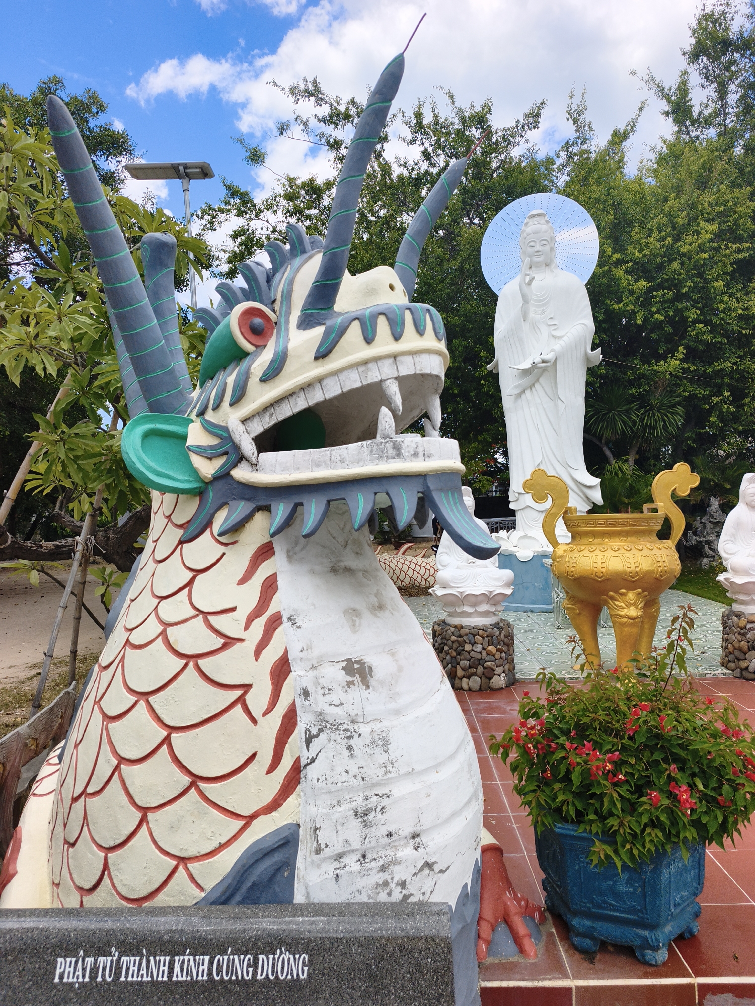 In Cam Ranh, Vietnam, a dragon statue with cream-colored scales outlined in red, blue accents, and green decorative elements stands in the foreground. Its mouth is open, revealing a white interior. Behind the dragon, a large white statue of a figure, likely a deity, with a halo is visible. A golden ornate urn with three legs and decorative handles sits to the right of the deity statue. A blue planter filled with green foliage and small red flowers is positioned in the lower right corner of the image, resting on a terracotta-tiled surface. The background features lush green trees and a bright blue sky with scattered white clouds. A solar-powered lamp post is visible among the trees in the upper left of the frame. A stone plaque with white text, appearing to be Vietnamese, is situated at the bottom of the image.