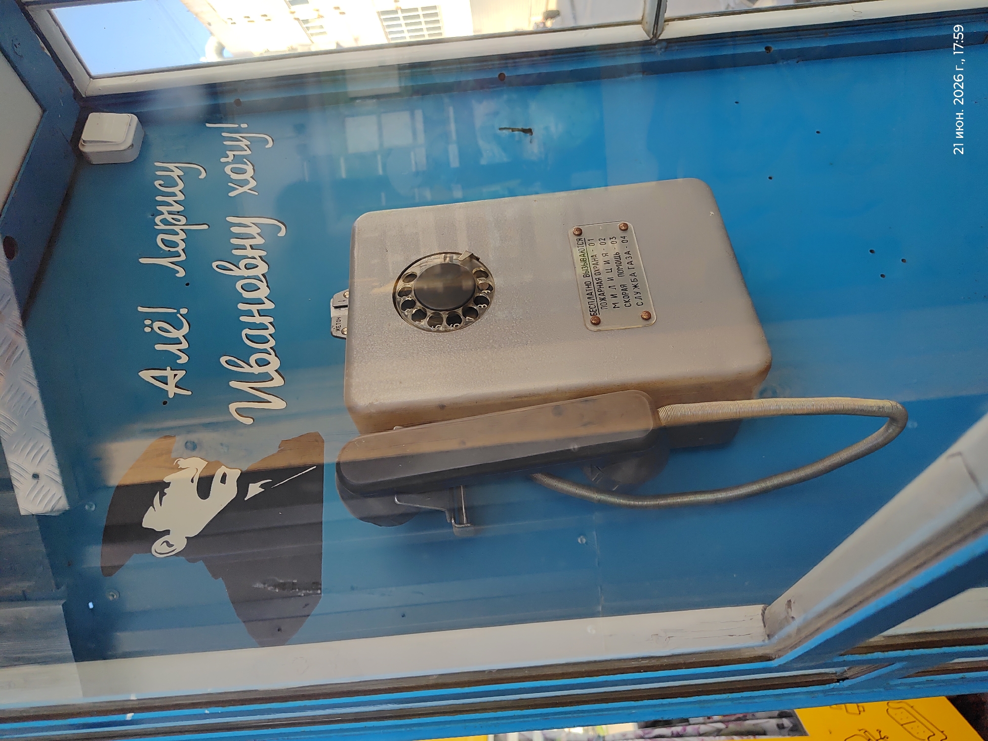 A public phone booth is depicted, painted a bright blue. On the back wall of the booth, a stencil of a man's profile in black and white is visible. To the right of the stencil, white cursive lettering reads, "Алё! Ларису Ивановну хочу!" which translates to "Hello! I want Larisa Ivanovna!". A black telephone receiver hangs on a metal hook to the left of a gray rotary dial phone. A small metal plate below the phone has white text listing emergency numbers: "БЕСПЛАТНО ВЫЗЫВАЮТСЯ" (FREE CALLS), "ПОЖАРНАЯ ОХРАНА - 01" (FIRE DEPARTMENT - 01), "М Л И Ц И Я - 02" (POLICE - 02), "СКОРАЯ ПОМОЩЬ - 03" (AMBULANCE - 03), and "СЛУЖБА ГАЗА - 04" (GAS SERVICE - 04). The phone booth appears to be in an urban environment, with a building visible through the glass window on the right. A date stamp at the bottom right corner of the image reads "21 июн. 2026 г., 17:59".