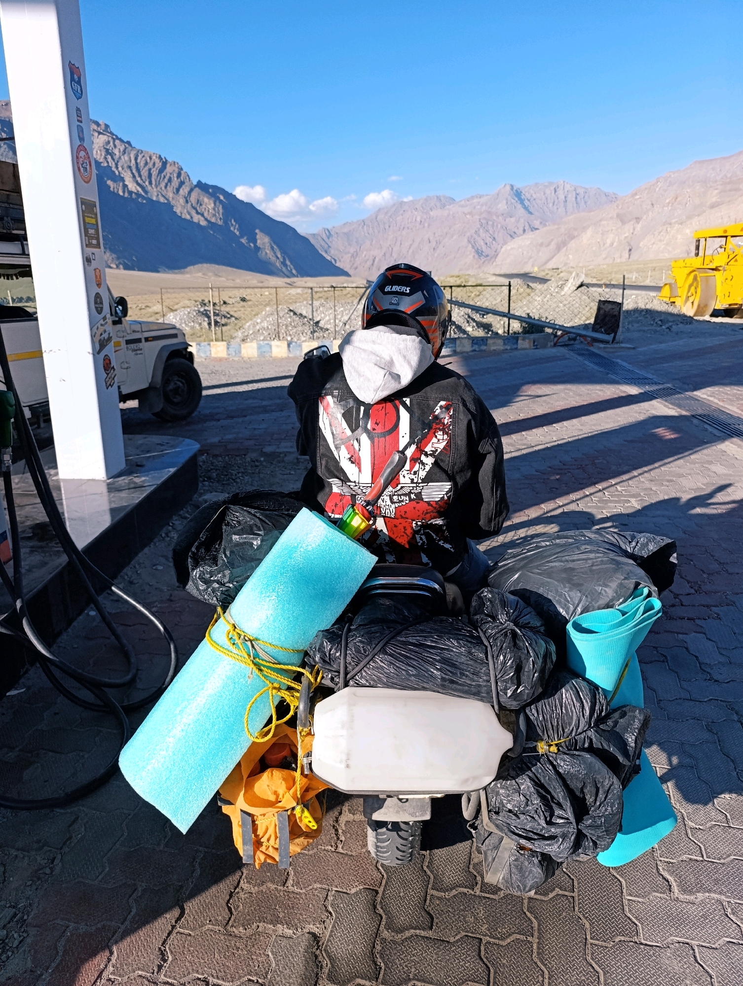 The image depicts a motorcycle packed with camping gear, including sleeping mats, bags, and a helmet, parked at a fuel station. The rider is seated on the motorcycle, facing away from the camera, wearing a black jacket and a helmet. In the background, mountains are visible under a clear blue sky. A white jeep and a yellow construction vehicle are also present at the fuel station. The scene is set in Pibiting, India, as indicated by the presence of a fuel pump with "Pibiting" visible on a sticker. The ground is paved with interlocking tiles, and shadows suggest it is daytime.