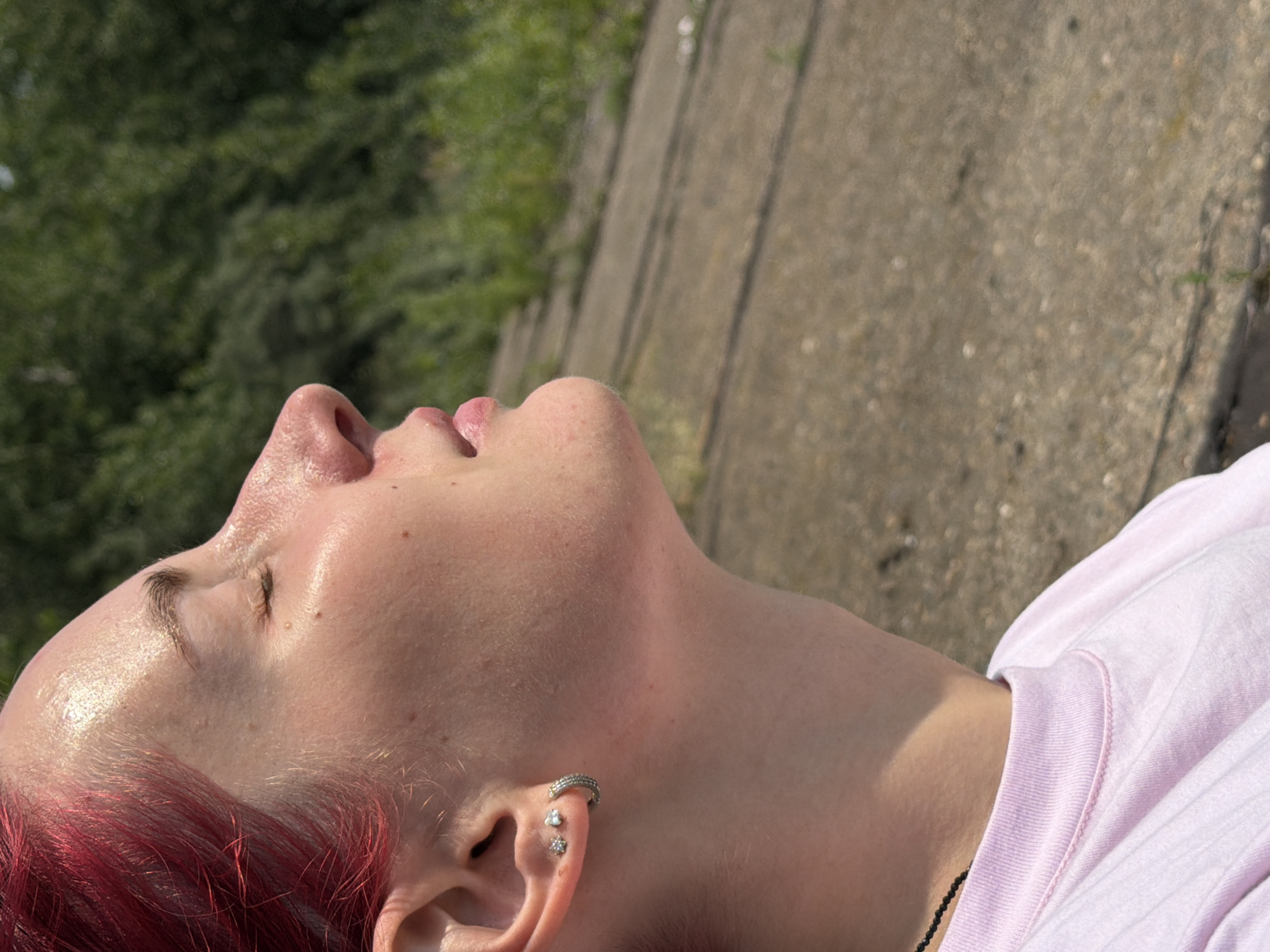 The image shows a close-up profile view of a person with vibrant red hair and closed eyes, facing upwards towards sunlight. They are wearing a pale pink t-shirt and a thin black beaded necklace. The person has small earrings in their left ear, one a stud with a star-like shape and the other a hoop earring. Their skin appears slightly shiny, likely from sweat or sunscreen.
The background consists of a textured concrete surface with some patches of green vegetation and what appears to be a railroad track or a similar linear path. Behind that, there is a dense, out-of-focus green foliage, suggesting an outdoor natural environment.
While the image is not definitively identifiable as being in Chaikovsky, Russia, the overall setting of a paved area adjacent to natural greenery is common in many urban and suburban environments. Without further context or specific landmarks, pinpointing the exact location within Chaikovsky is not possible based solely on this image.