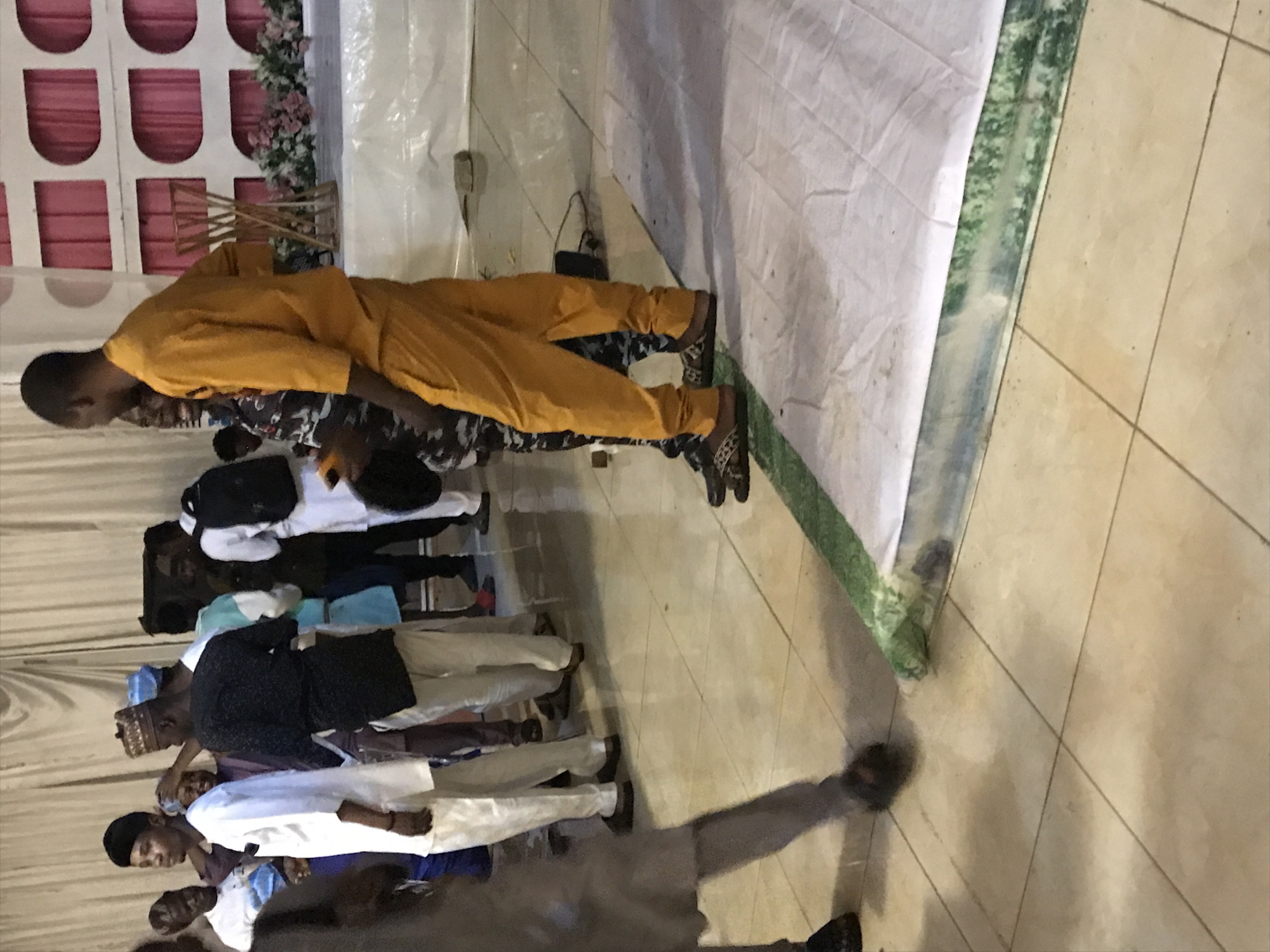 A group of people are standing indoors, likely at an event in Oko Erin, Nigeria. One person is wearing an orange jumpsuit. Several others are wearing white shirts with dark pants or blue bottoms. A white fabric covering is in the background. The floor has a tile pattern.